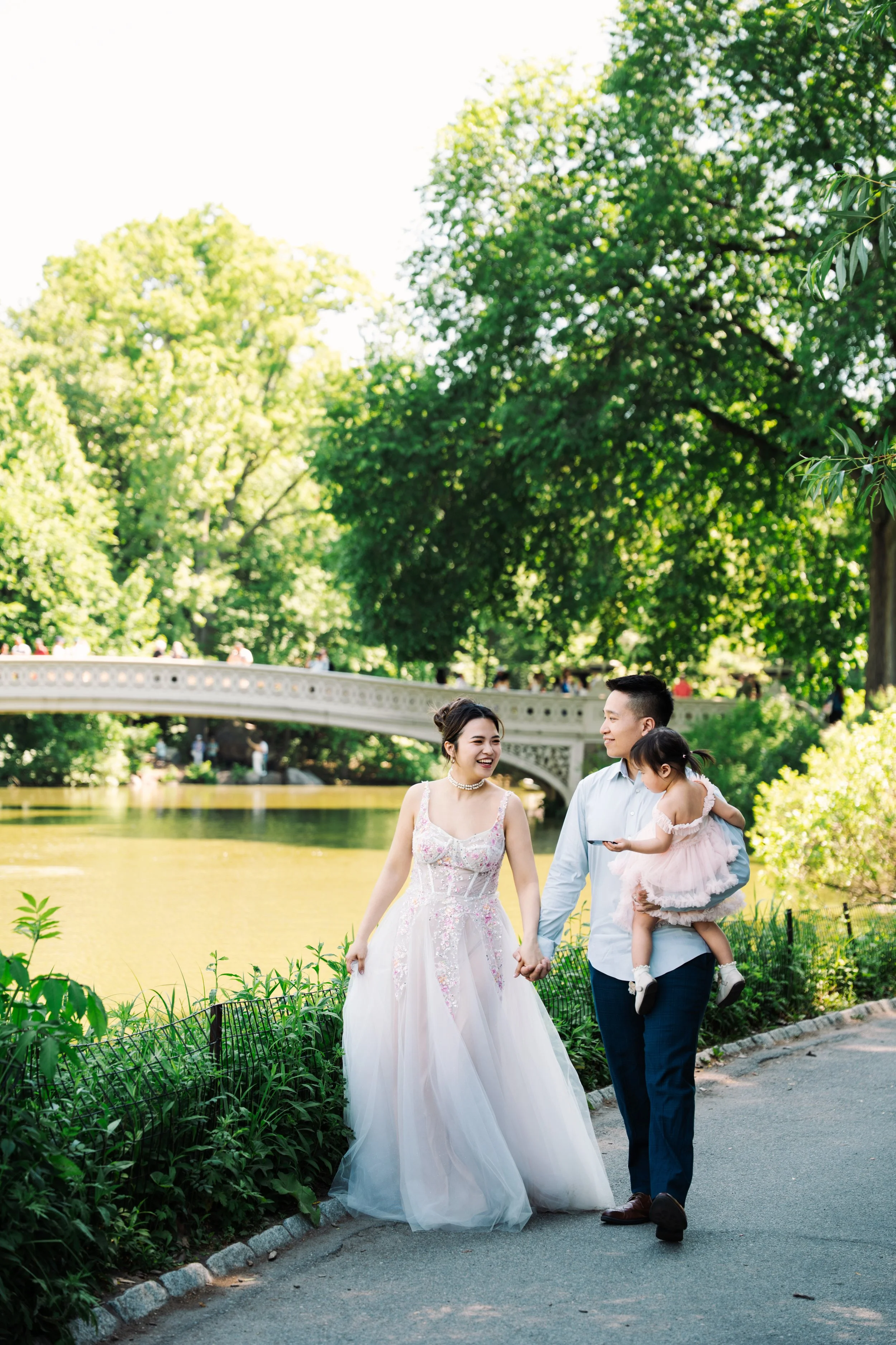A joyful family walking in a park. The woman is in a wedding dress, and the man is carrying a young girl in a pink dress. They are holding hands and smiling, with a bridge and green trees in the background.