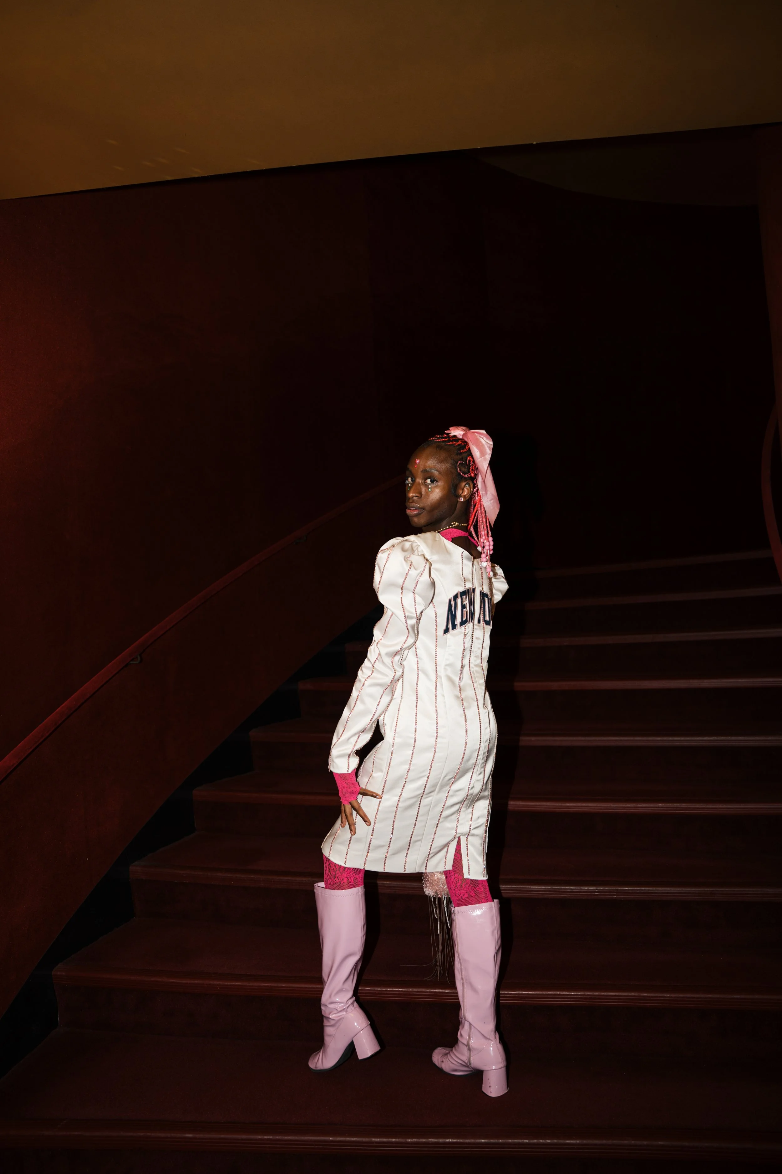 A woman dressed in a white and pink streetwear outfit with thigh-high pink boots, standing on a staircase in a dark environment with no visible background details.