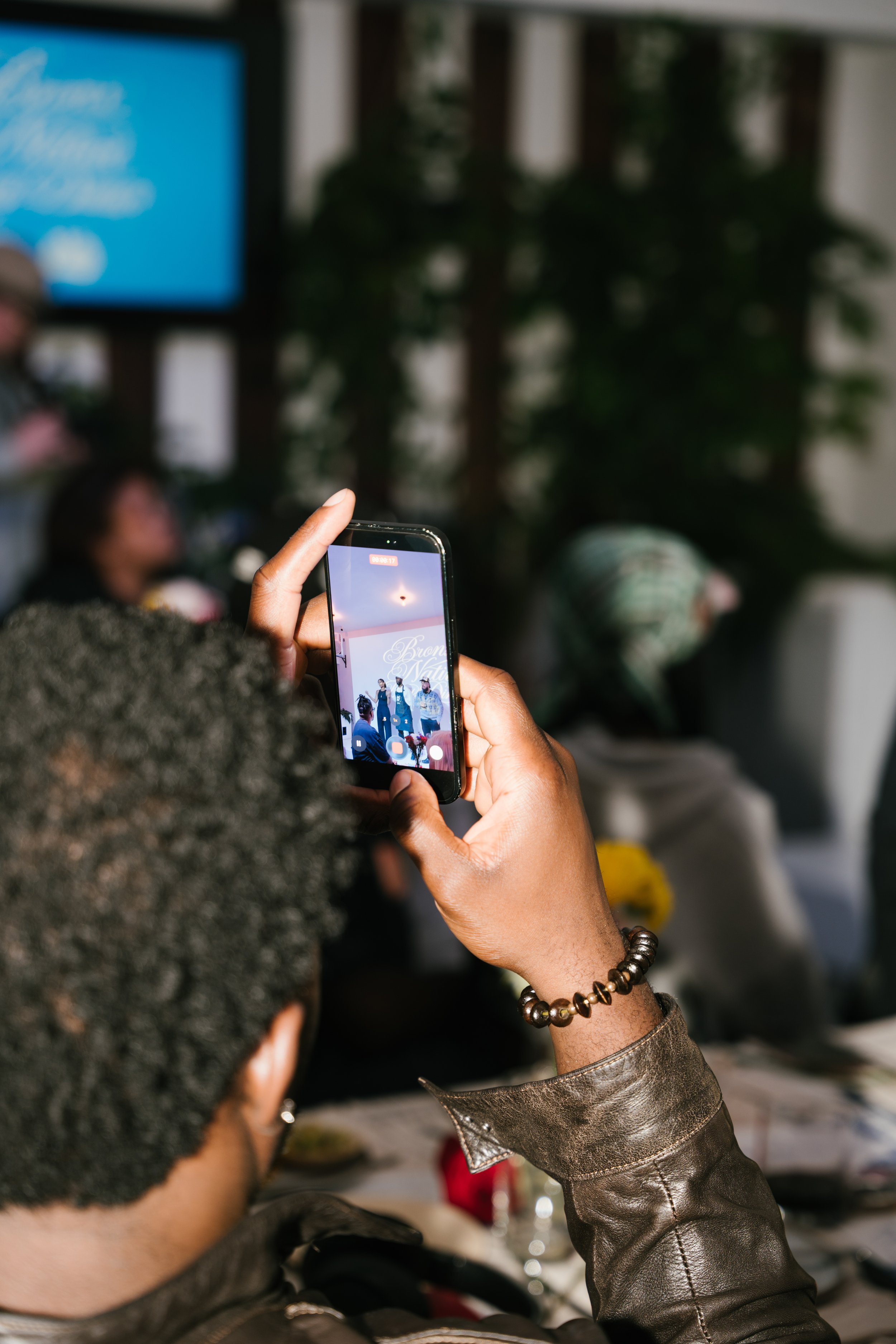 Person holding a smartphone capturing an image of a band performing on stage, with a blurred background including a woman and some greenery.