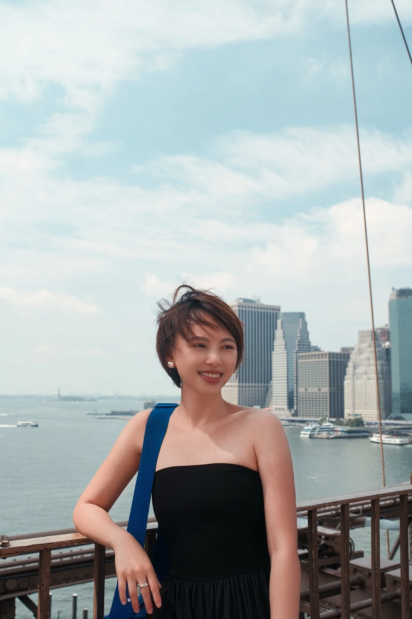 A woman with short brown hair standing on a bridge with a city skyline and river in the background, smiling, wearing a black strapless top and a blue bag over her shoulder.
