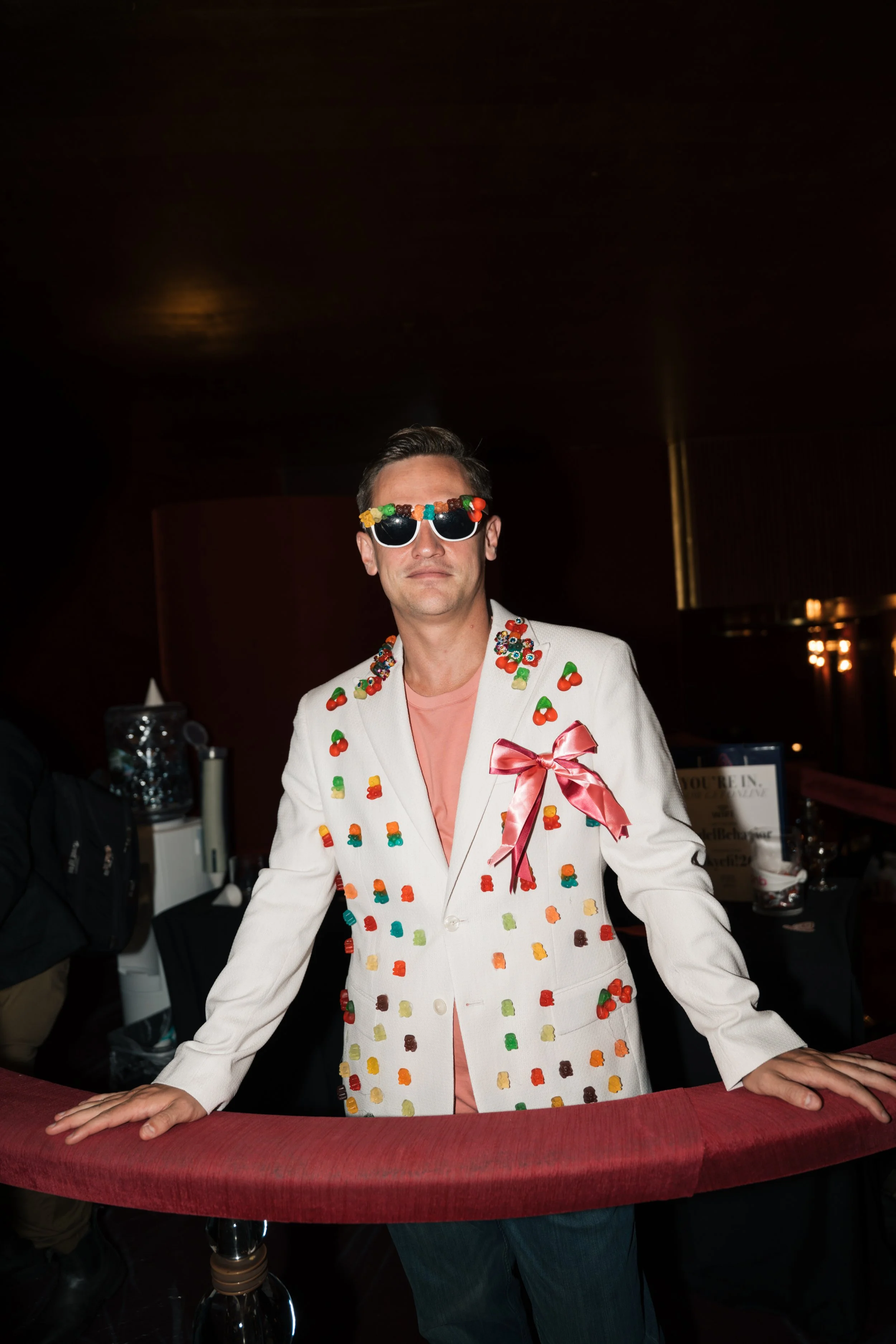 Man wearing a white suit decorated with toy candies and chocolates, pink shirt, pink bow on suit, and sunglasses with gummy bear decorations, standing at a table in a dimly lit event space.