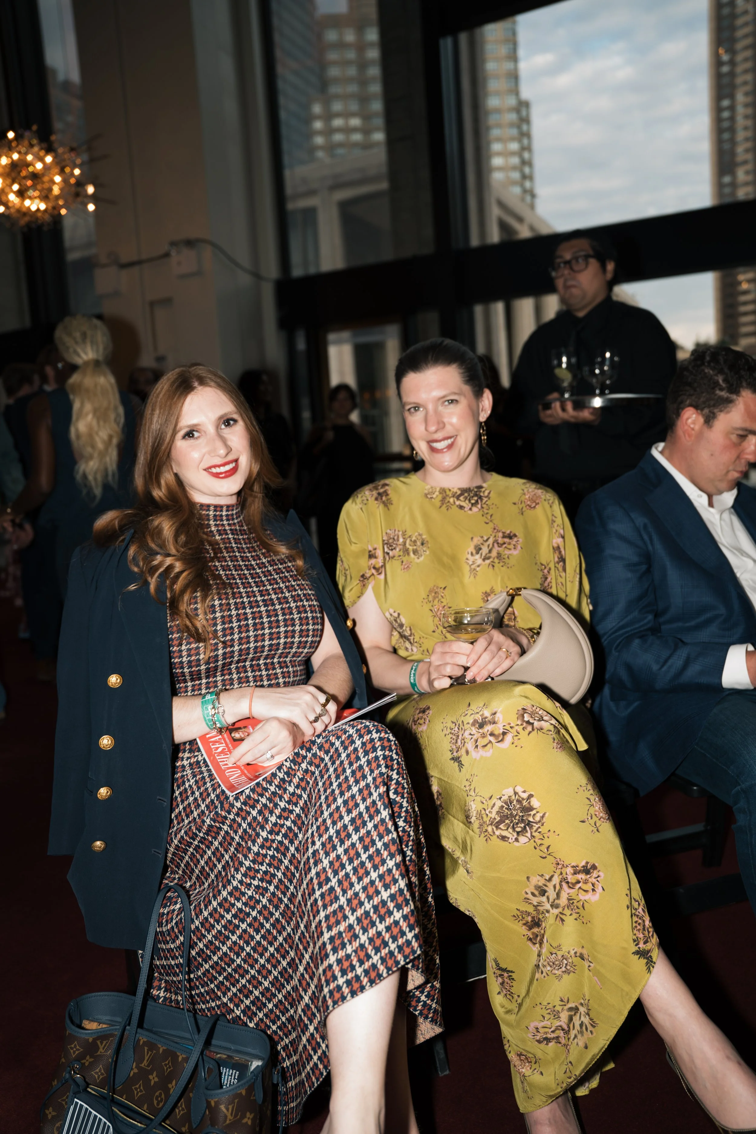 Two women sitting at a table during a social event in a tall building with large windows, cityscape view, and other guests in the background. One woman has long red hair, wearing a patterned dress and navy blazer, holding a magazine and smiling. The 