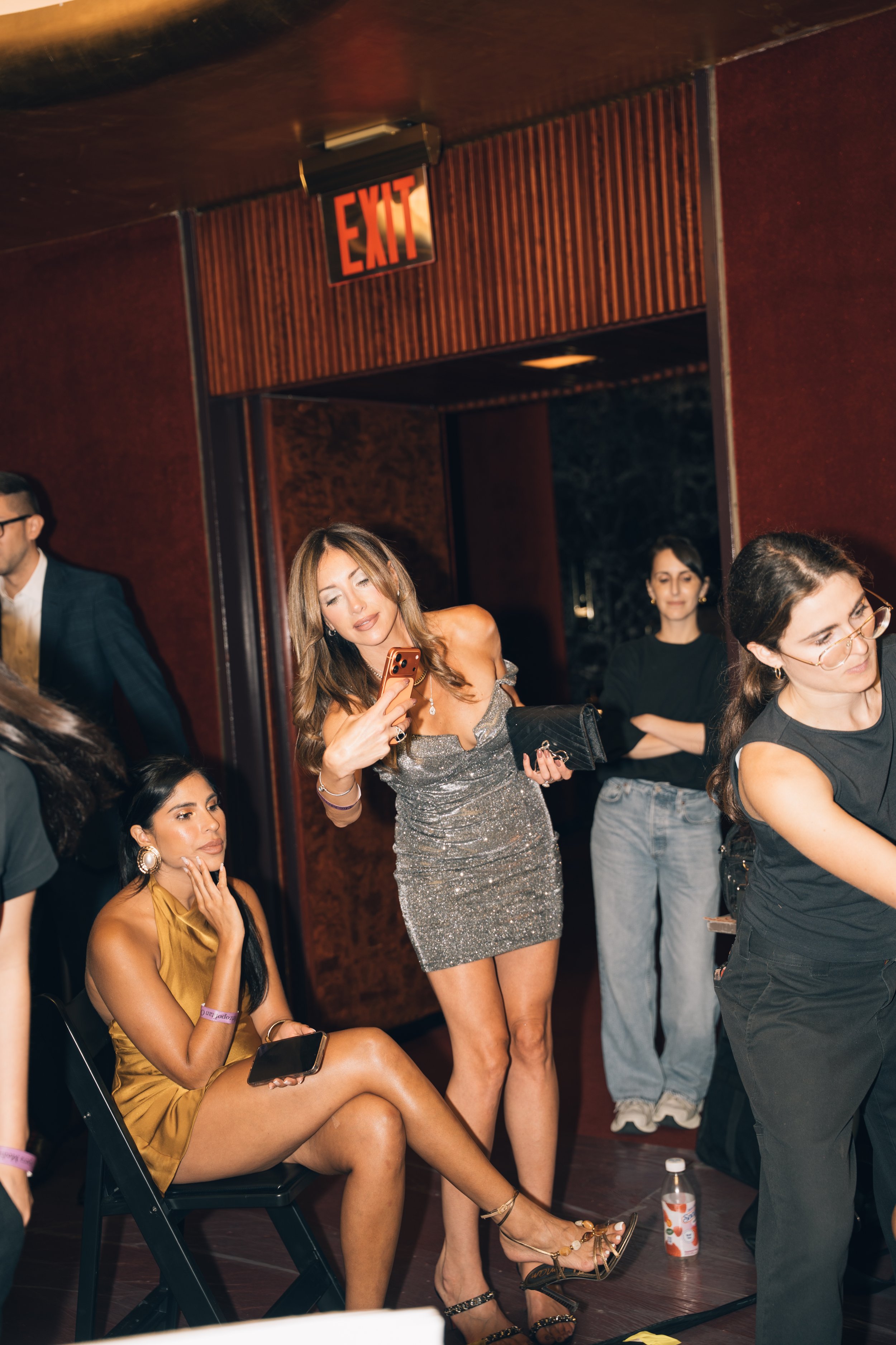 A group of women at an indoor event. One woman in a gold dress is sitting on a folding chair, holding a smartphone, with her legs crossed and wearing high heels. Another woman in a sparkly silver dress is standing nearby, looking at her phone, with h