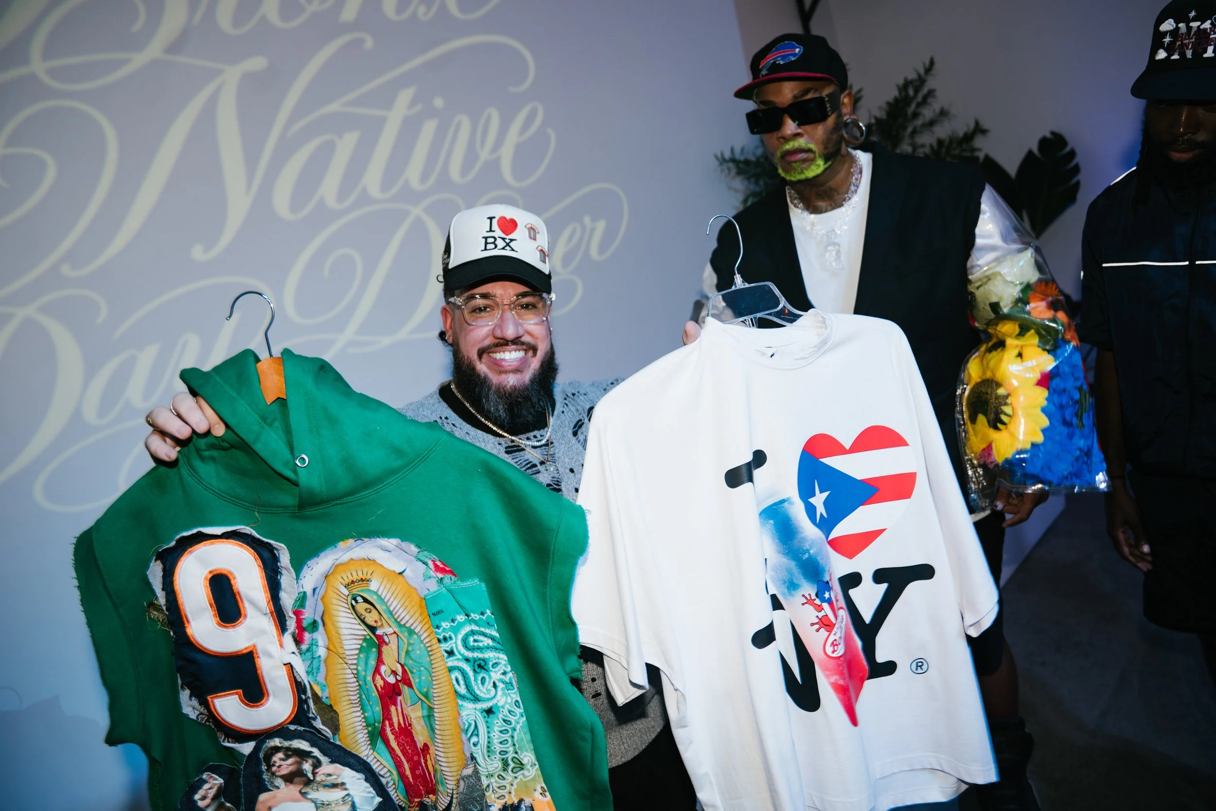 Two men holding hangers with clothing, one smiling and wearing a green jacket with patches including a '93' and a portrait, and a baseball cap with 'I ❤️ BX', the other wearing black sunglasses, a black cap with colorful designs, and a black vest, wi