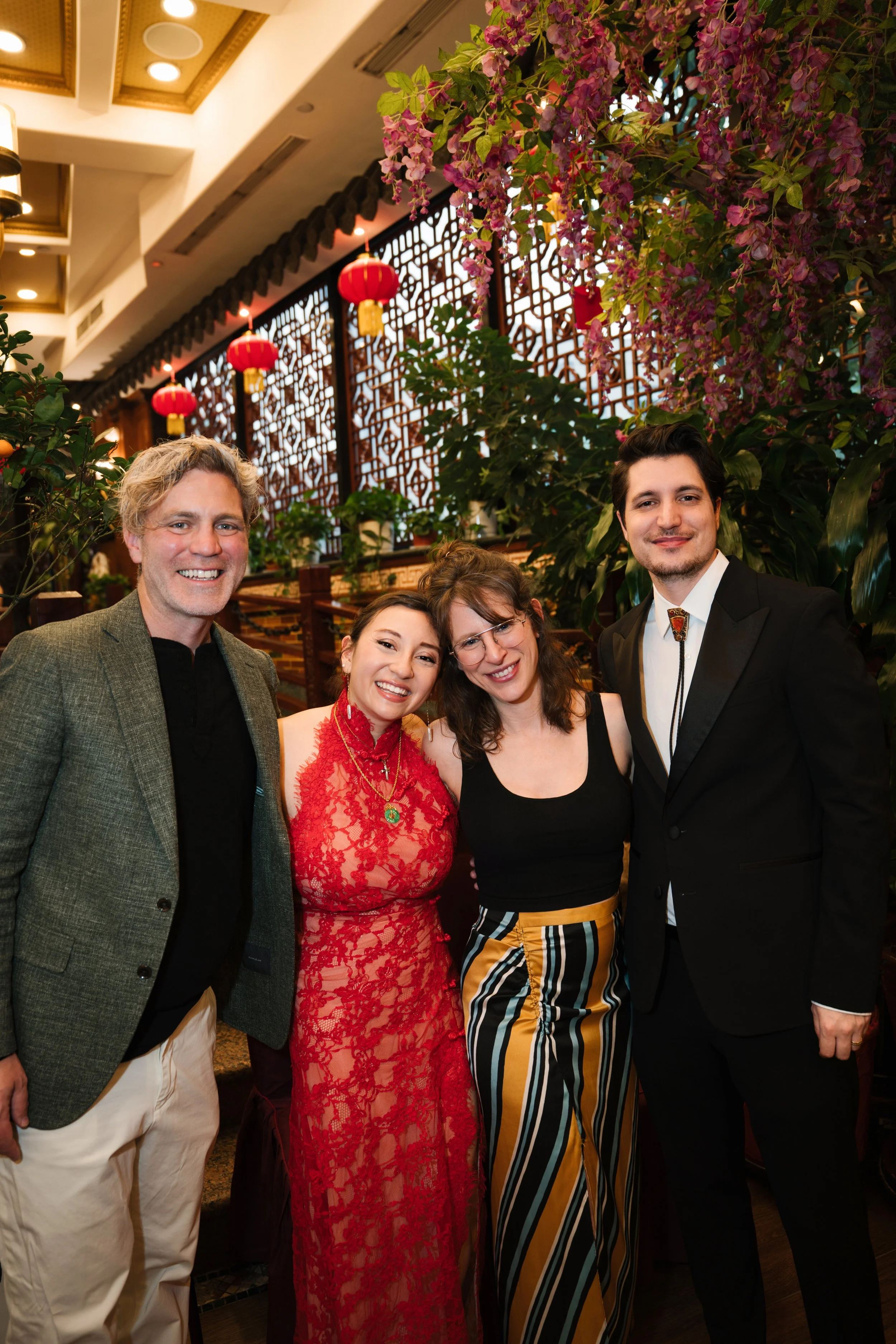 Group of five people smiling and posing for a photo in a decorated indoor setting with pink flowers and red lanterns hanging from the ceiling.