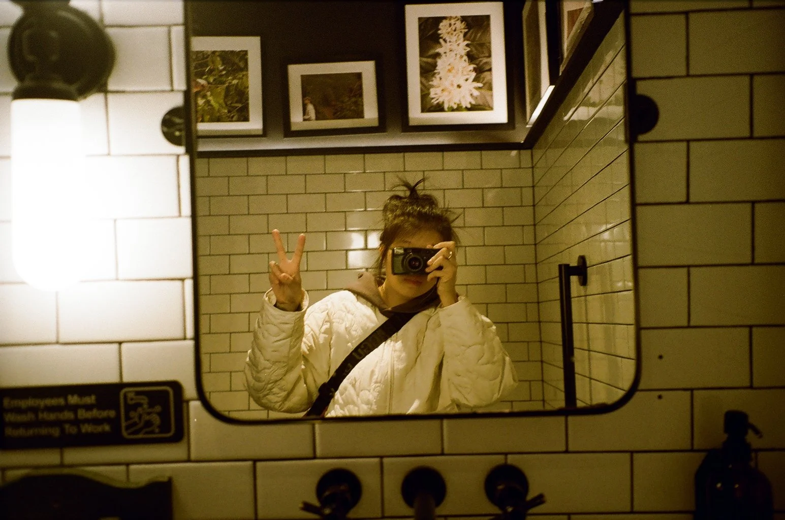 A person with a messy bun hairstyle taking a mirror selfie in a bathroom. They are wearing a white quilted jacket, holding a camera with one hand, and flashing a peace sign with the other. The bathroom has white subway tiles on the walls, framed pictures above, and a black metal towel holder.