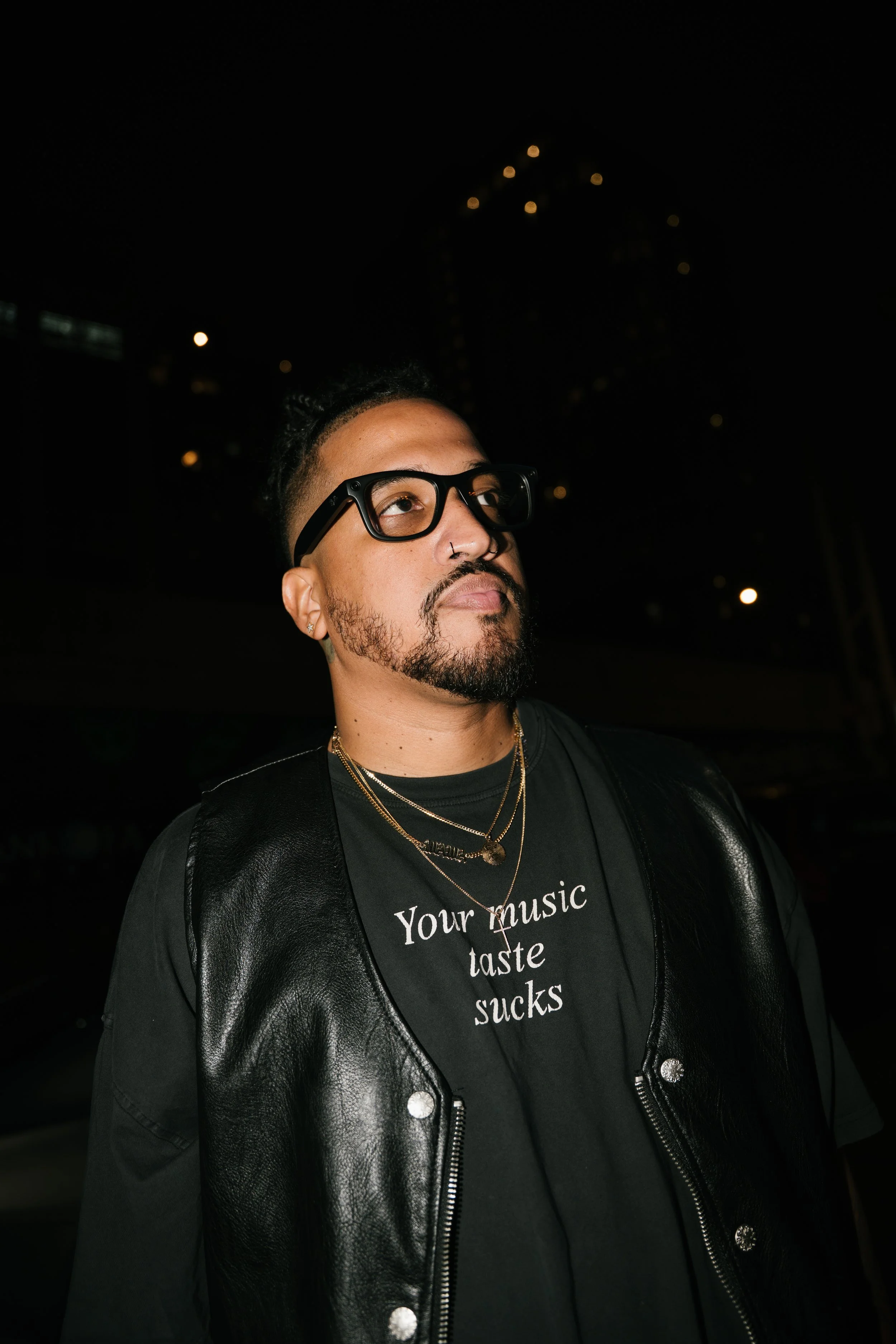 A man with short dark hair, glasses, and a nose piercing, wearing a black leather vest over a black t-shirt that says "Your music taste sucks," at night with city lights in the background.
