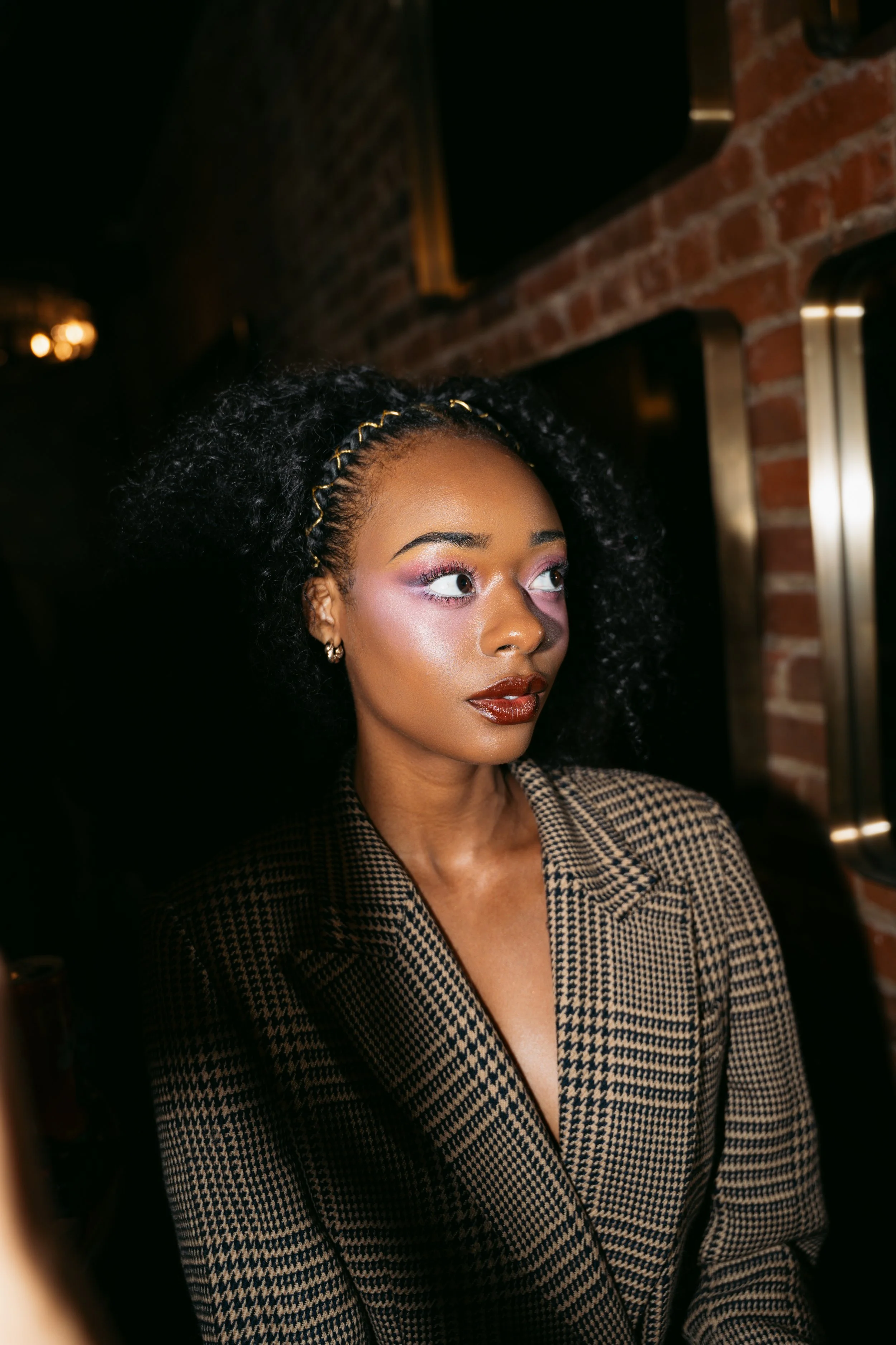 Portrait of a woman with curly black hair, wearing a patterned blazer, with makeup and earrings, in an indoor setting with a brick wall background.