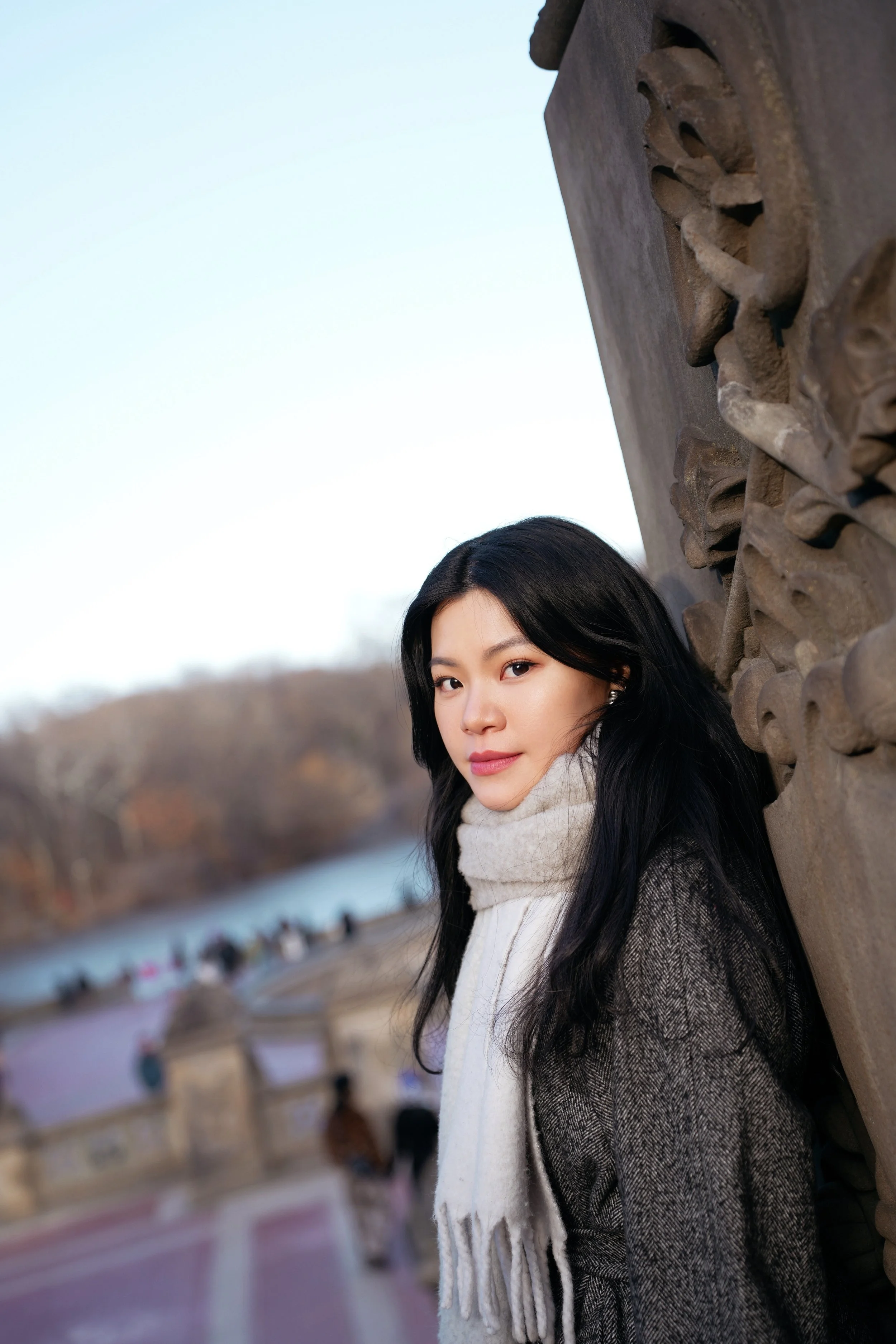 A young woman with long black hair, wearing a dark coat and a cream-colored scarf, standing outdoors next to an ornate stone wall with a river and trees in the background.