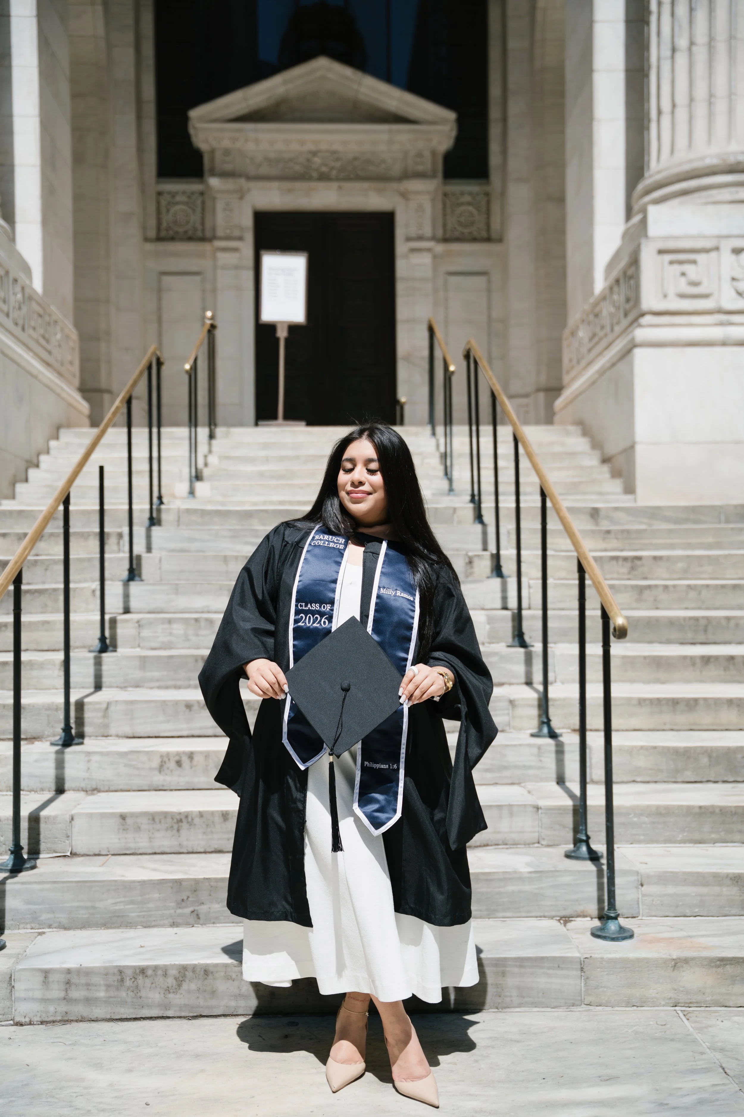 A young woman in a graduation gown and cap standing at the top of a stone staircase outside a historic building, holding her diploma and smiling.