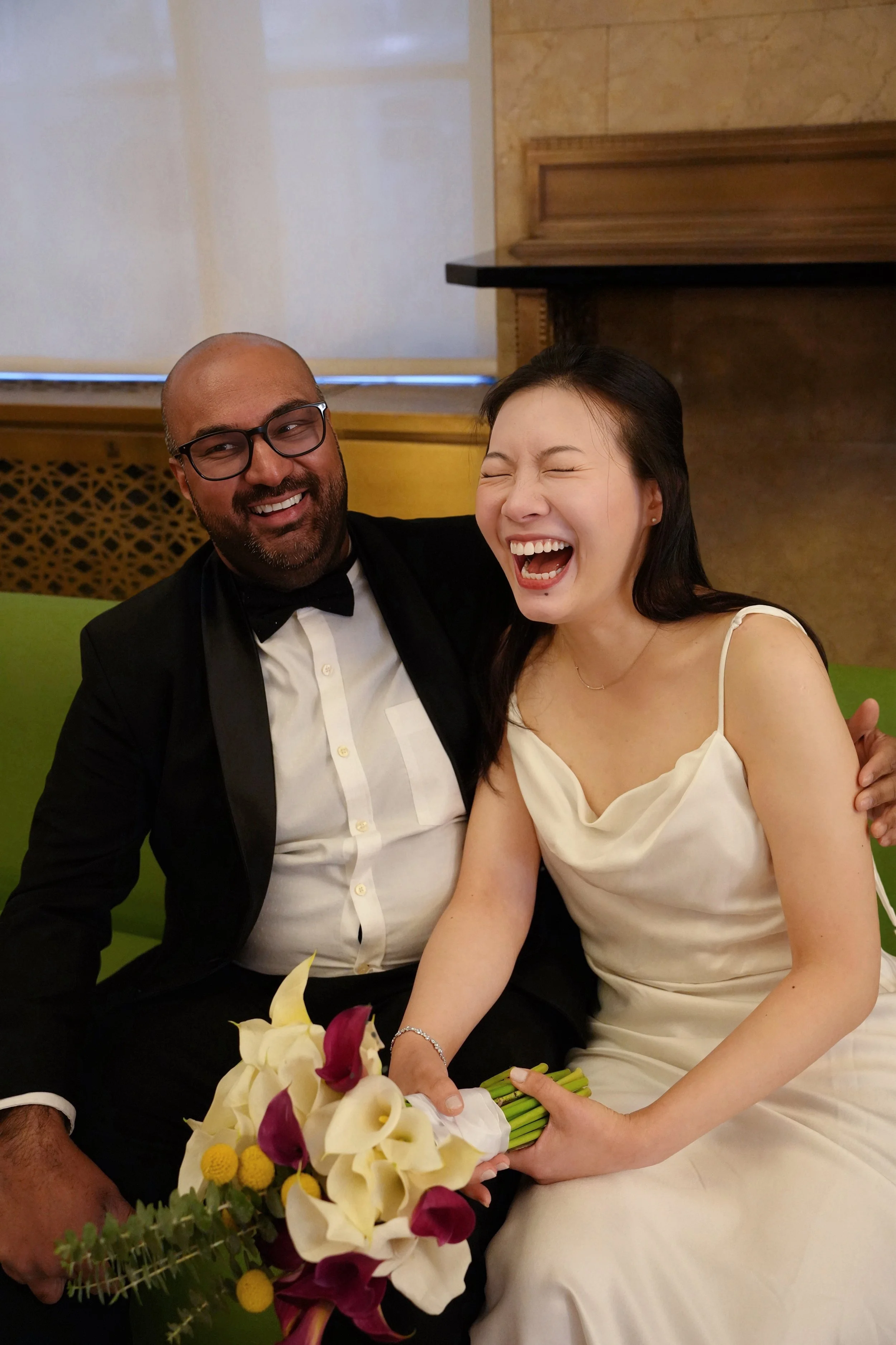 A joyful couple at a formal event, the man in a black tuxedo and glasses, the woman in an elegant white dress, holding a bouquet of flowers, both laughing.