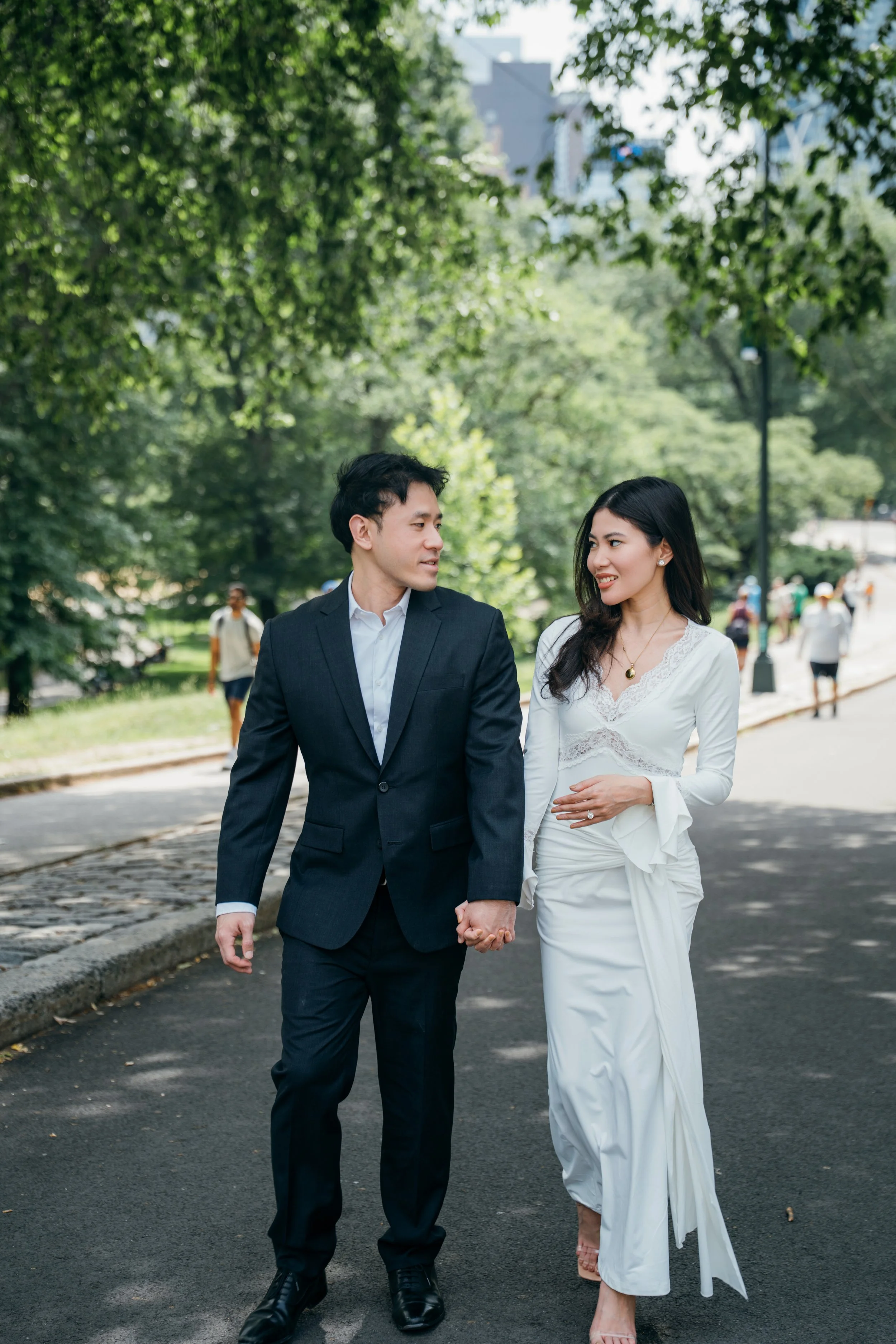 A couple walking hand in hand in a park, holding hands, with greenery and other park visitors in the background. The man is wearing a dark suit and the woman is wearing a white dress.