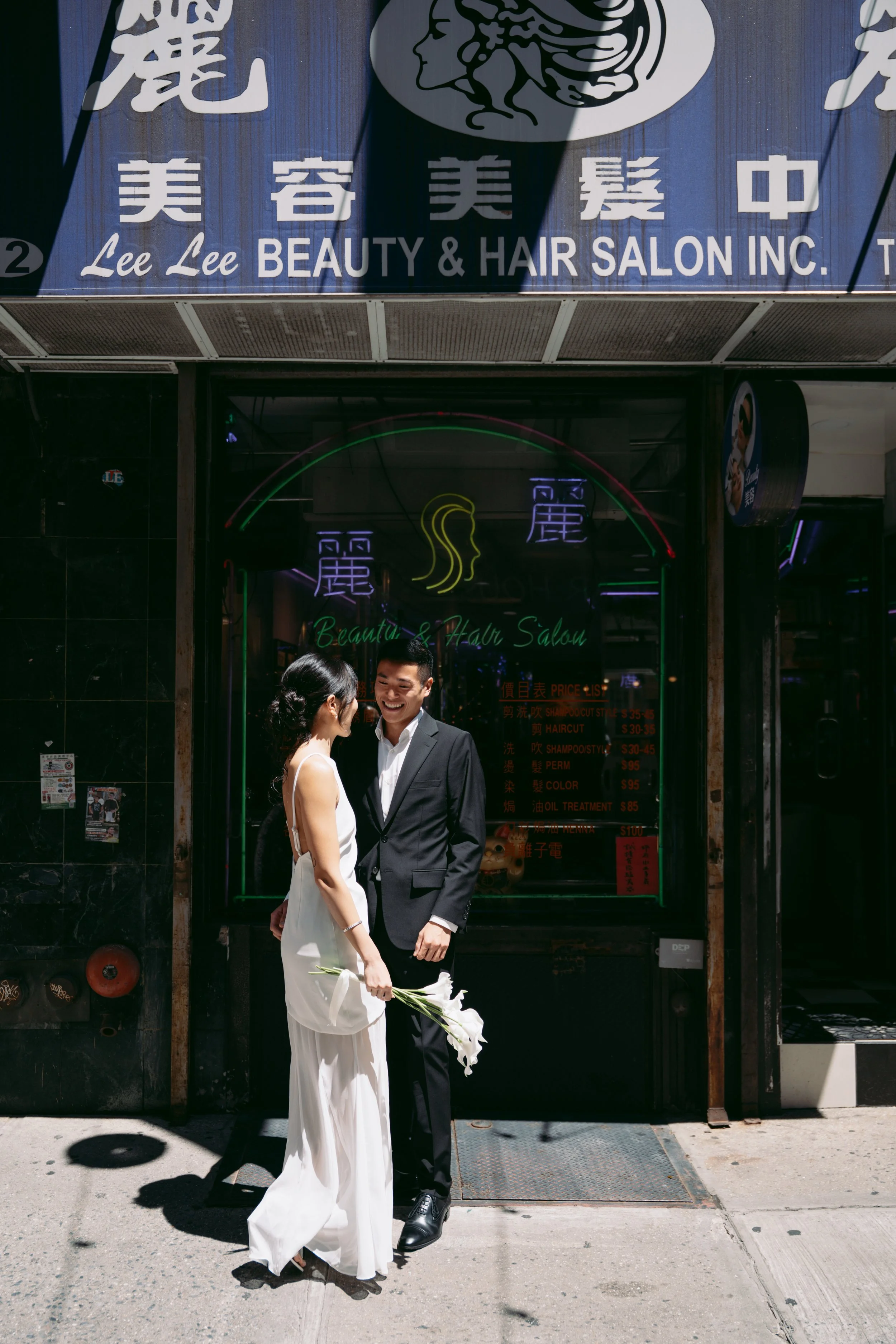 A bride and groom smiling and holding hands outside a beauty and hair salon entrance. The bride wears a white dress and holds a bouquet of white flowers, while the groom is dressed in a dark suit with a white shirt. The salon sign above features Chin