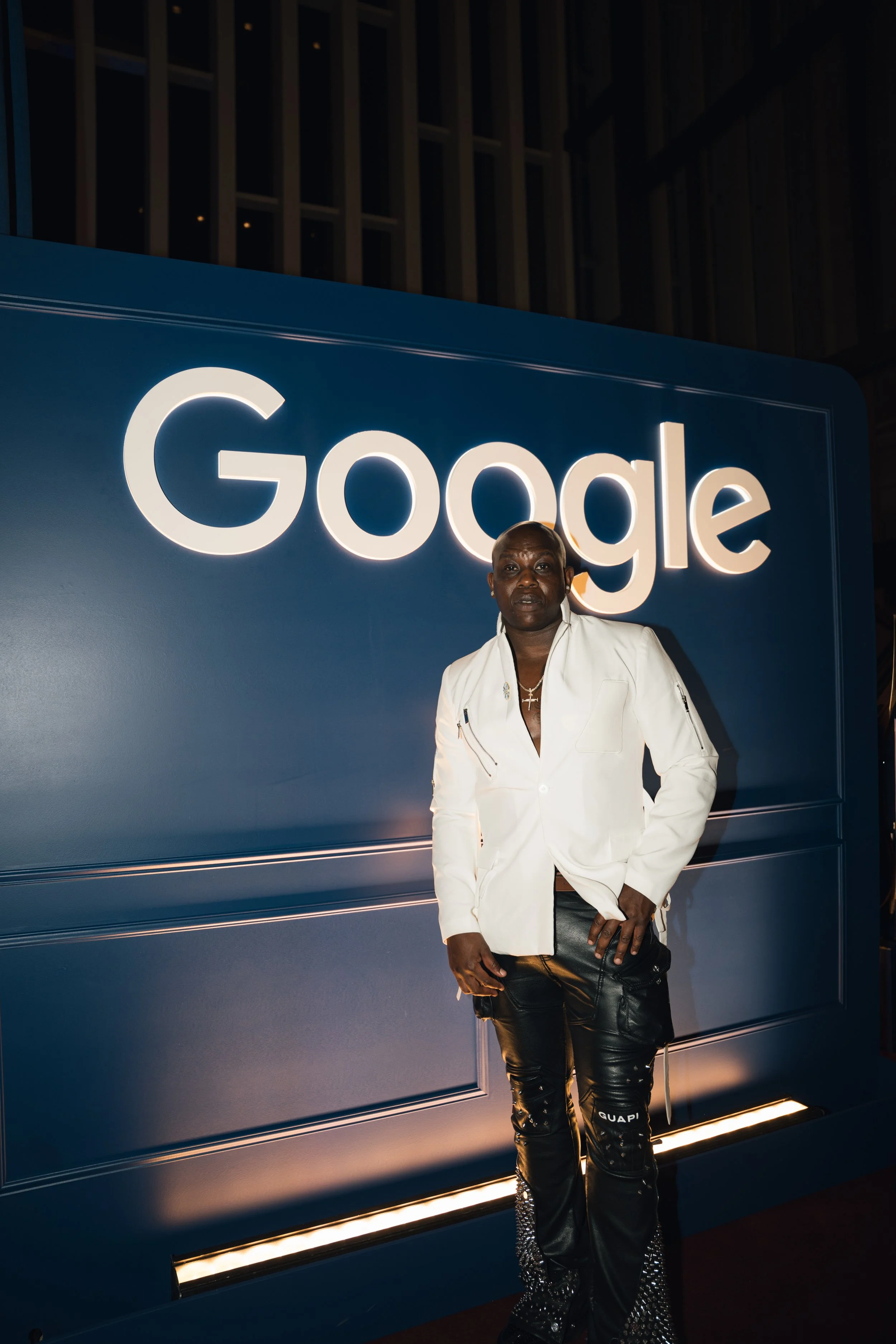 A person standing in front of a large illuminated Google sign at night, wearing a white jacket and black leather pants.