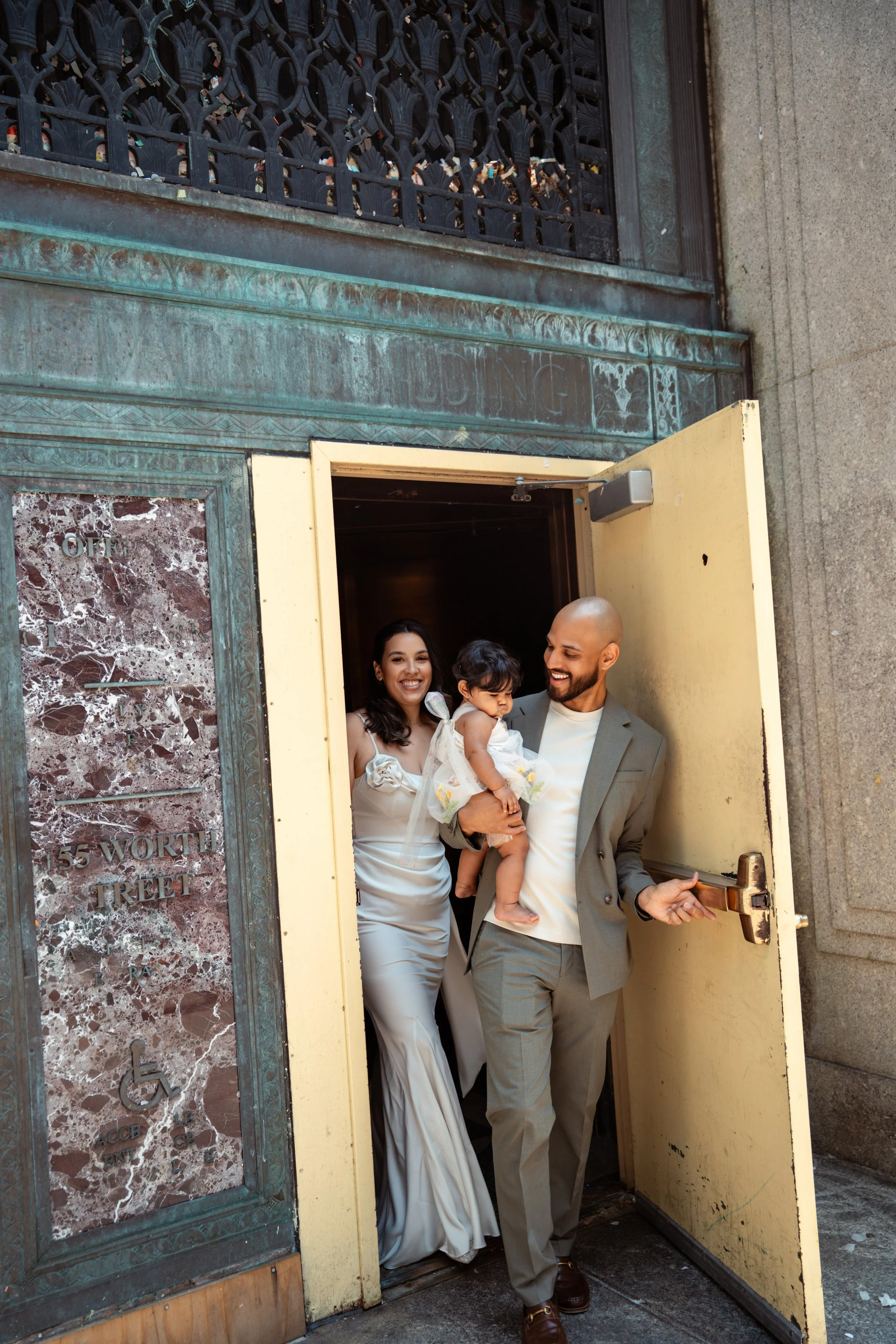 A family of three, two adults and a young child, exiting a door on the street. They are smiling and look happy, with the woman and child smiling at the camera and the man looking at the woman. The woman is in a white dress, the man is in a gray suit,