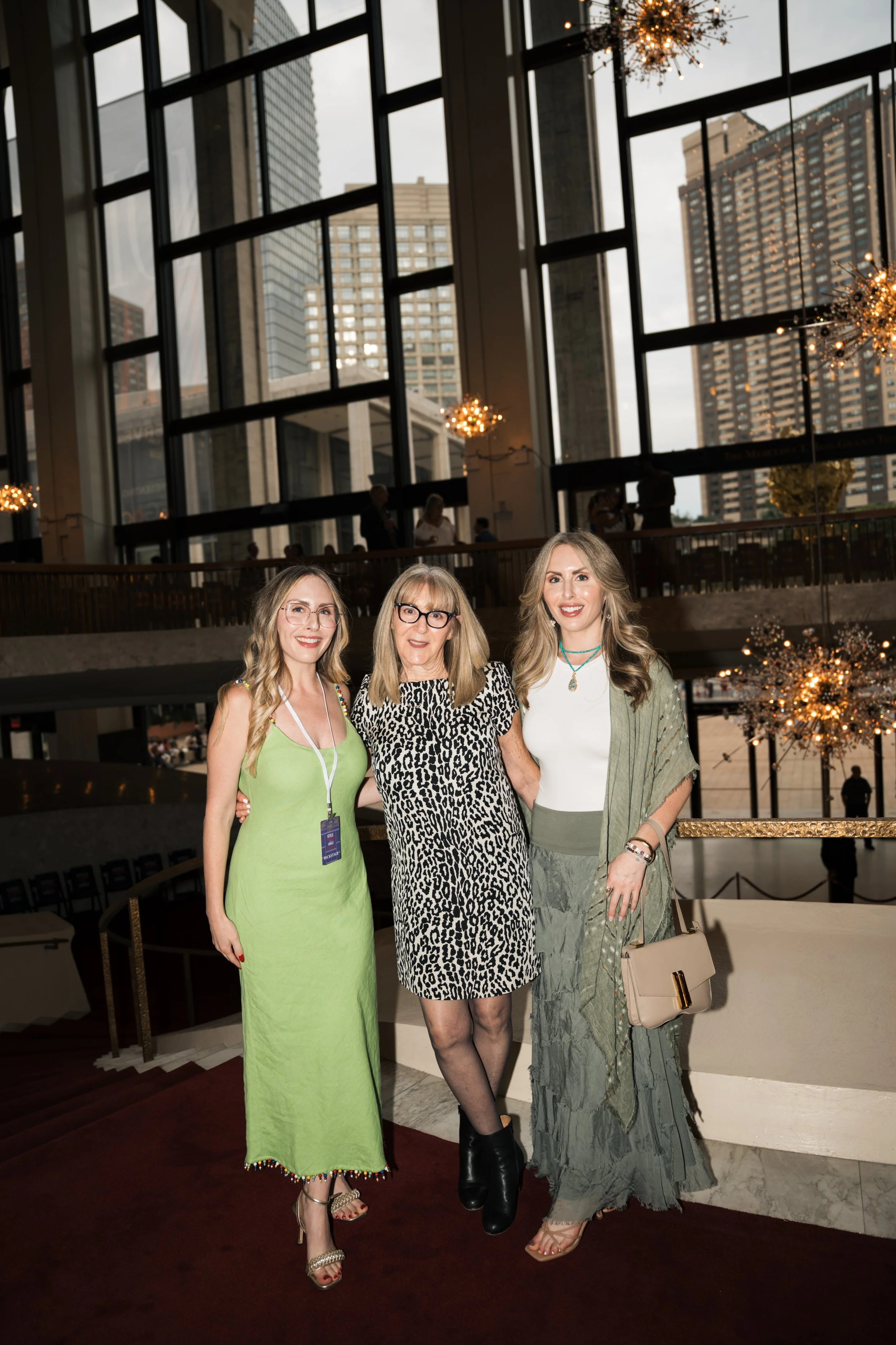 Three women, dressed in stylish outfits, standing together indoors at what appears to be a high-rise building with large windows and a view of tall city buildings. They are smiling and posing for a photo.
