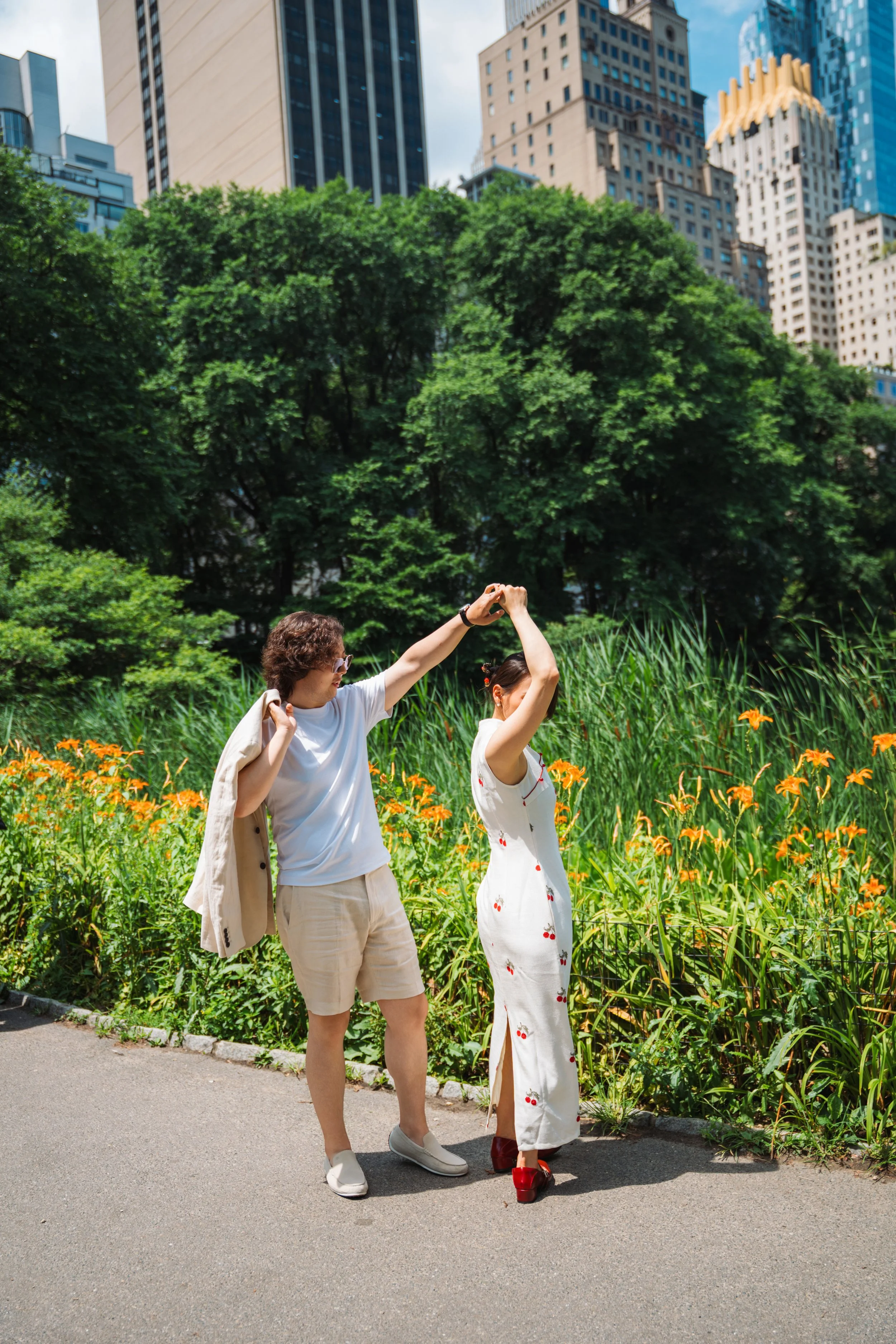 Two women are dancing and holding hands in an urban park with tall green trees and orange flowers in the foreground. One woman is wearing a beige blazer, white shirt, beige shorts, and white sneakers. The other woman is wearing a white dress with red
