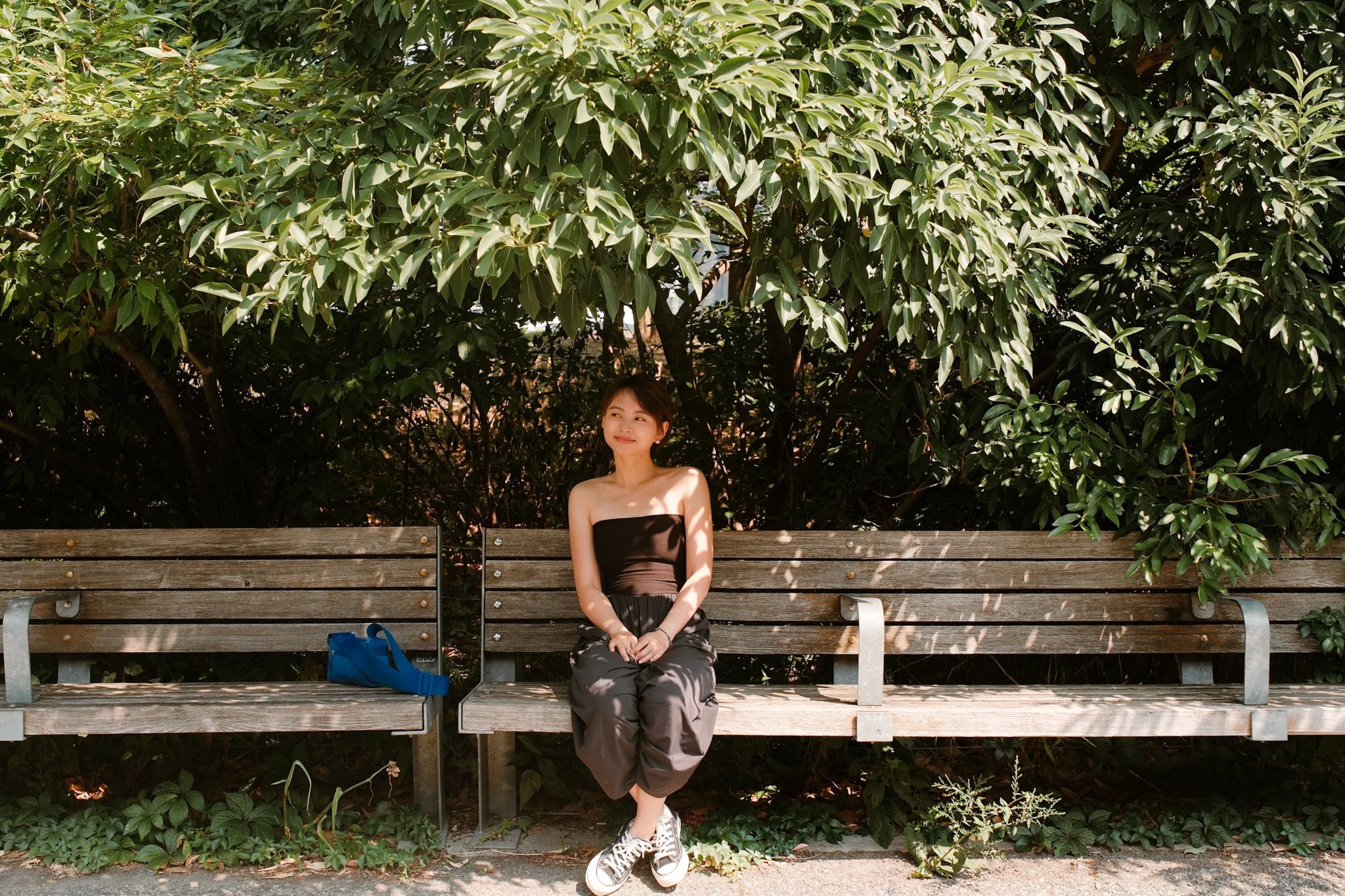 A young woman with short brown hair sitting on a park bench underneath green leafy trees, wearing a black strapless top, patterned pants, and white sneakers, with a blue bag placed on the bench beside her.