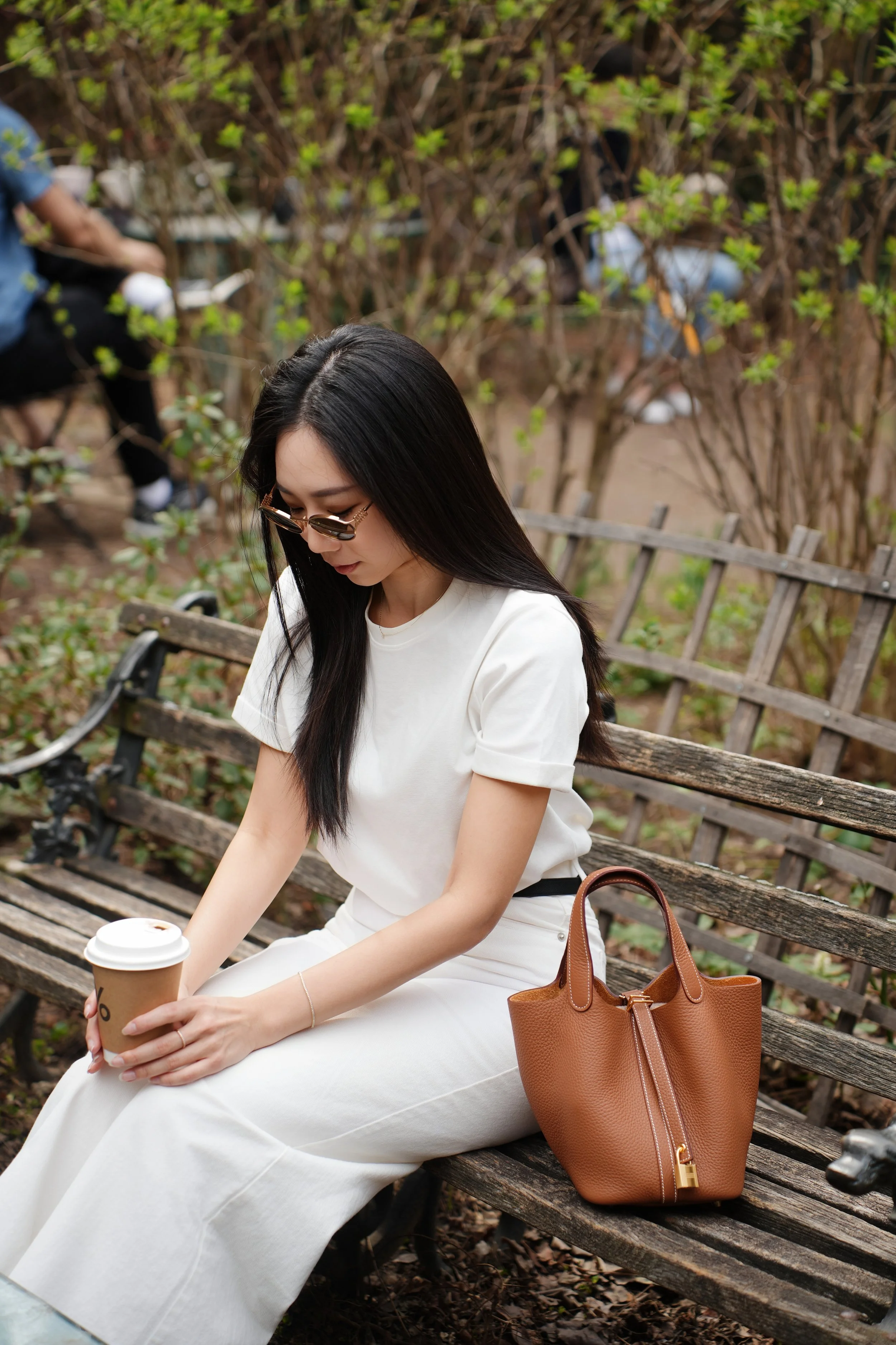 A woman with long black hair and sunglasses sitting on a wooden park bench, holding a coffee cup. She is wearing a white dress and a brown handbag is placed beside her on the bench. There are trees and bushes in the background.