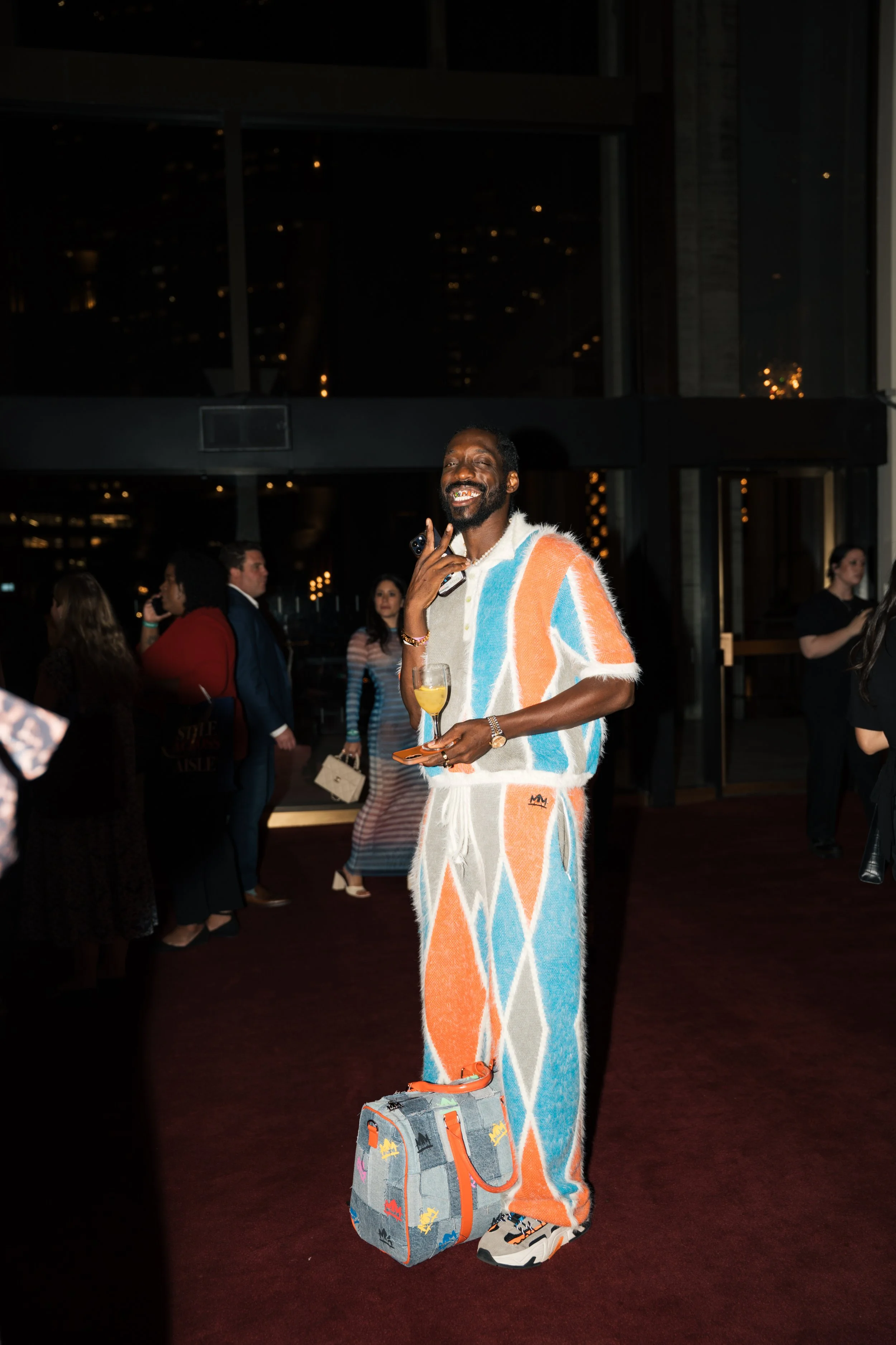 A man wearing a colorful geometric patterned jumpsuit, holding a drink, with a gray backpack featuring colorful moose motifs, at an indoor event at night with other people in the background.