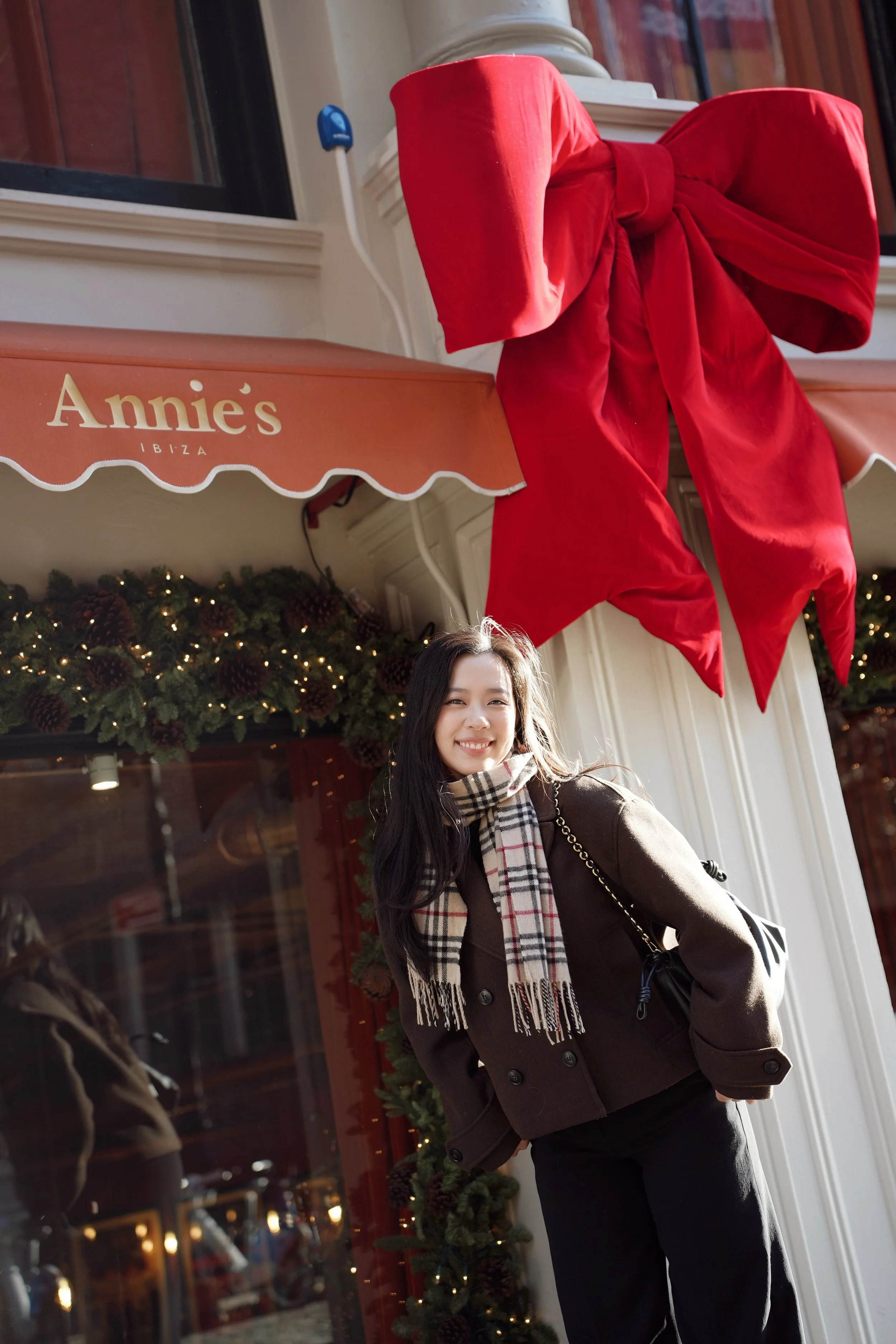 A smiling woman with long dark hair standing outside an Annie's Ibiza store, decorated with Christmas greenery and lights, wearing a brown coat, black pants, and a plaid scarf.