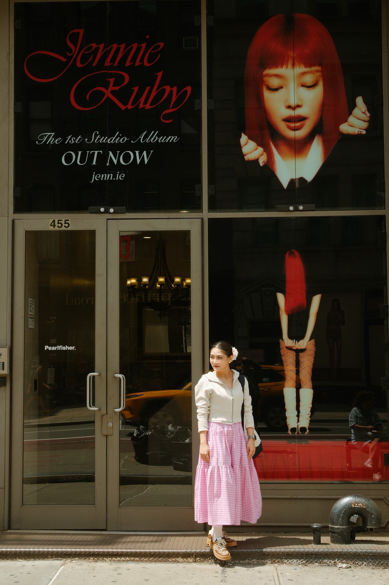 A woman wearing a pink and white checkered skirt, white jacket, and tan platform shoes stands in front of a store window. The window displays an advertisement for an album by Jennie from Blackpink, with a large image of Jennie with red hair and her e