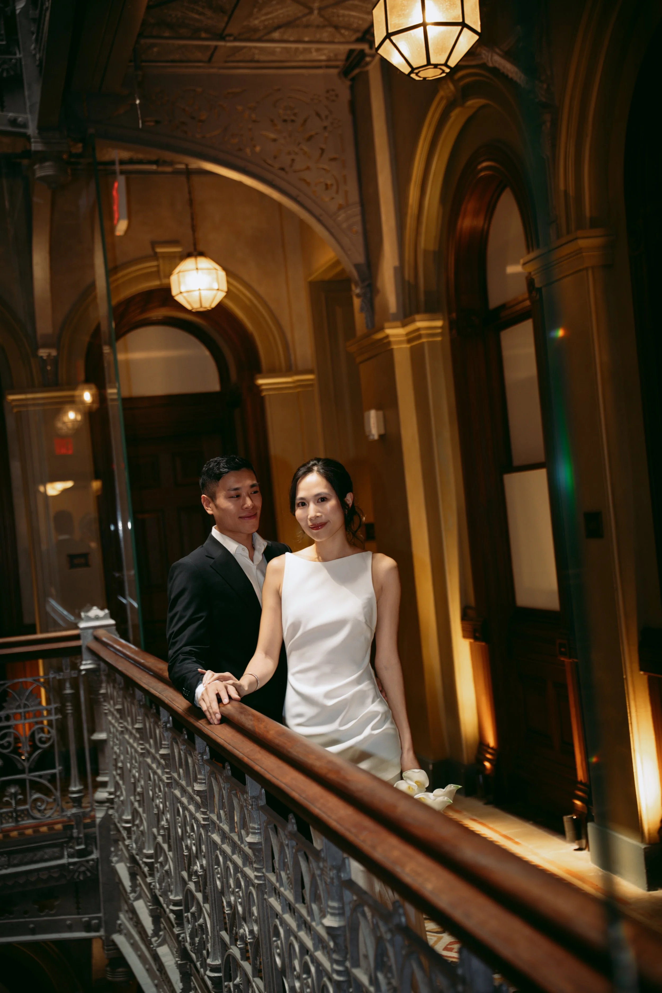 A couple dressed in formal attire, standing on a decorative balcony inside a building with ornate architectural details, wooden accents, and warm lighting from hanging chandeliers.