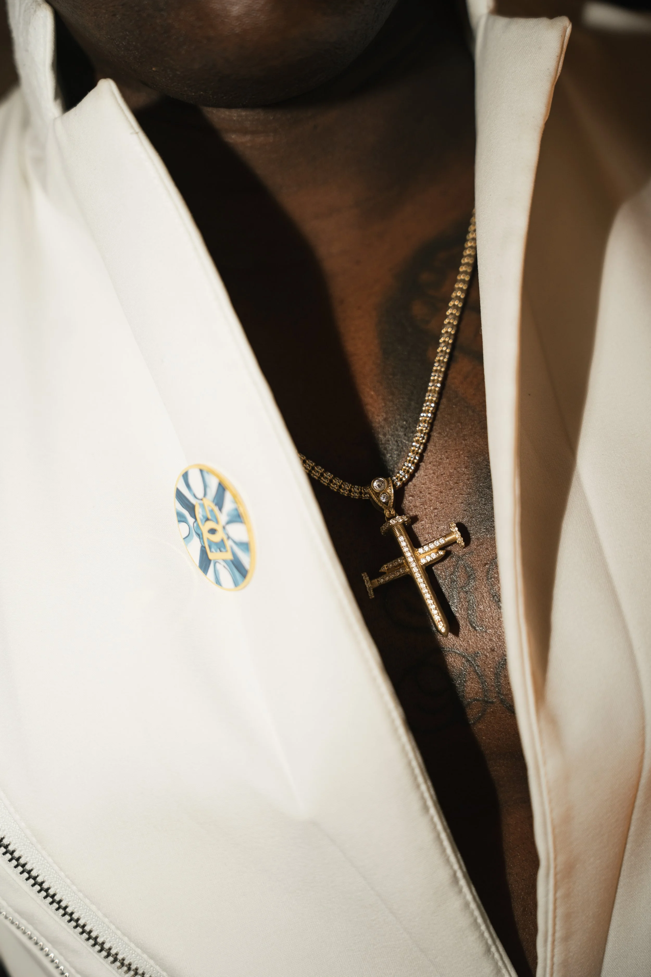 Close-up of a person's neck and chest with a gold chain necklace with a letter 'H' pendant, partially visible white jacket, and a gold and blue badge on the jacket.