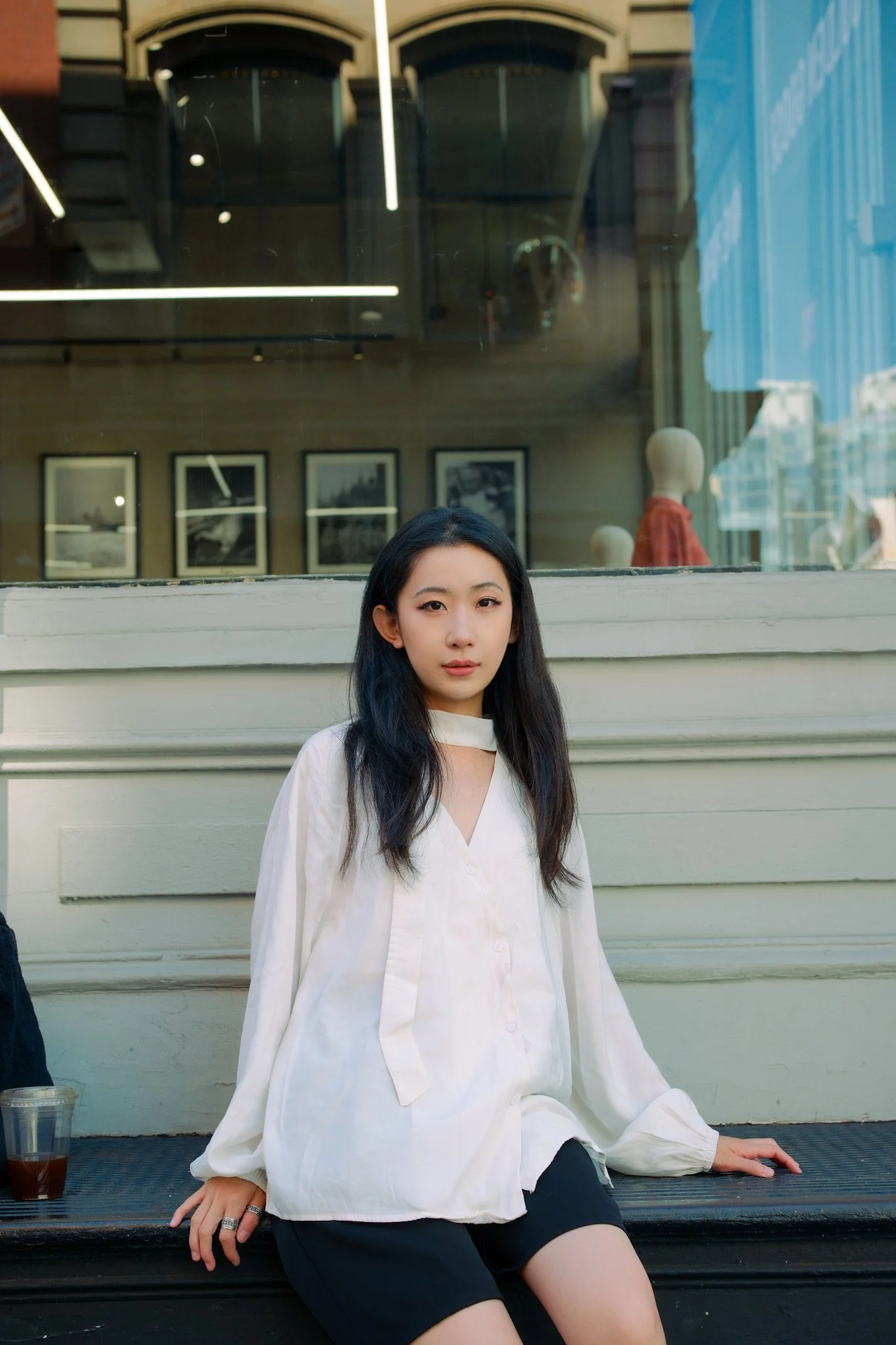 A young woman with long dark hair and fair skin sitting on a black bench outside a building. She is wearing a white blouse and black shorts, with a couple of rings on her fingers. There is a drink on the bench near her left hand. Behind her is a larg