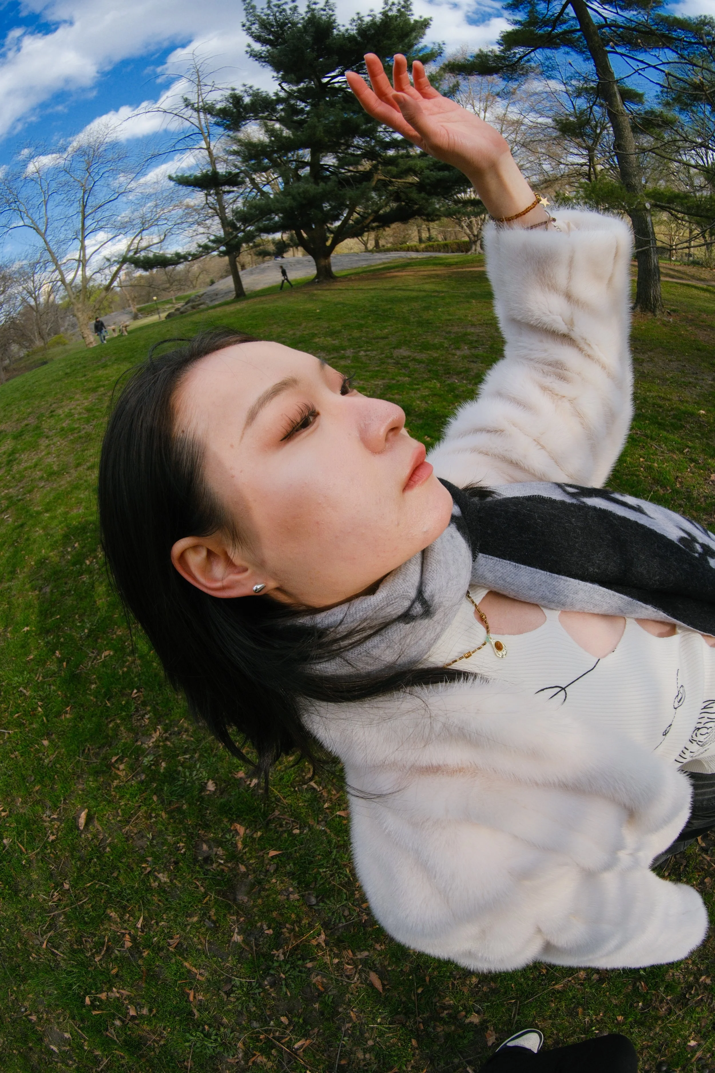 A woman lying on the grass in a park, with her eyes closed, wearing a furry white jacket, jewelry, and a gray hoodie, surrounded by trees and a blue sky with clouds.