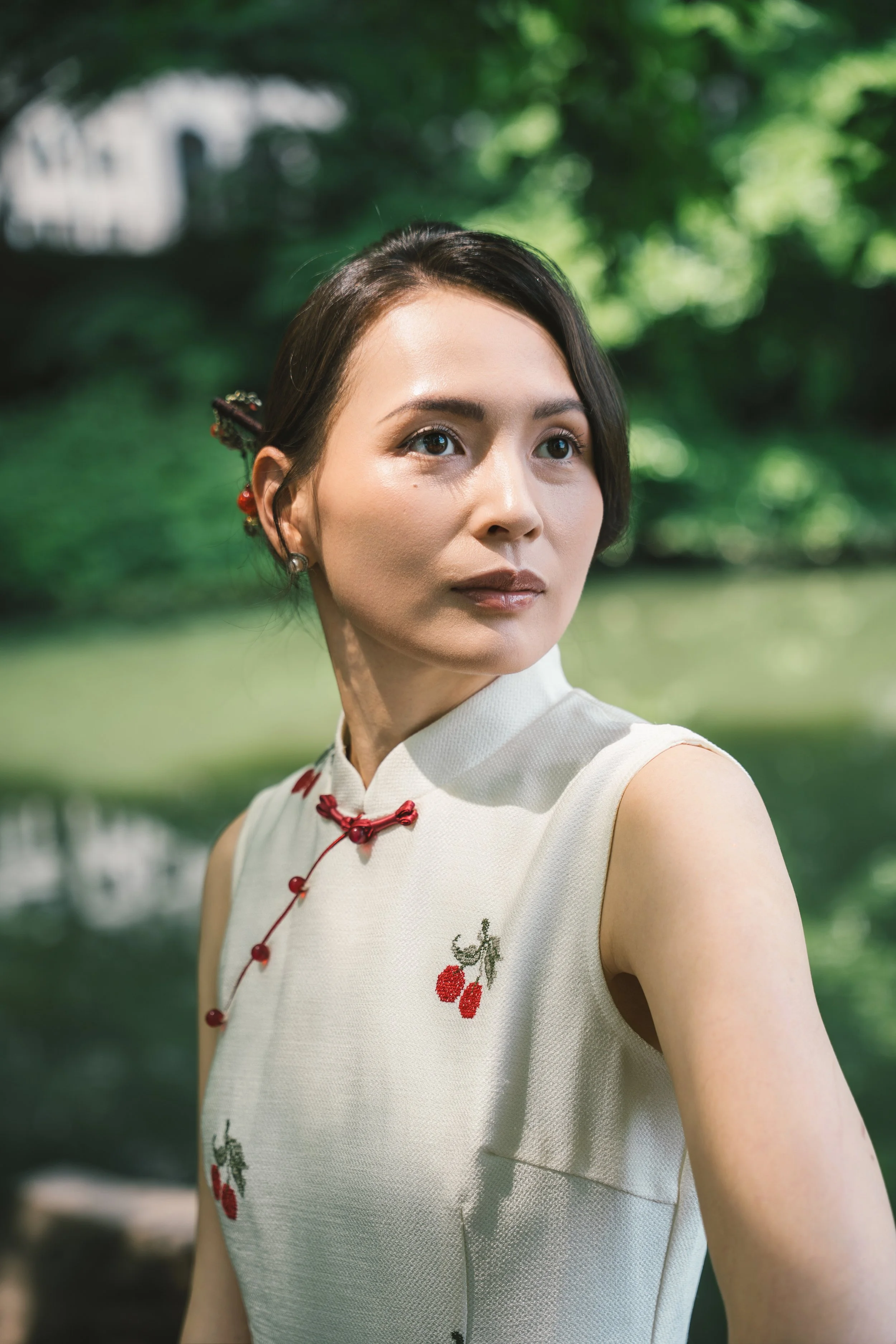 A woman with dark hair styled back, wearing earrings and a sleeveless white top with cherry embroidery, stands outdoors with green trees and sunlight in the background.