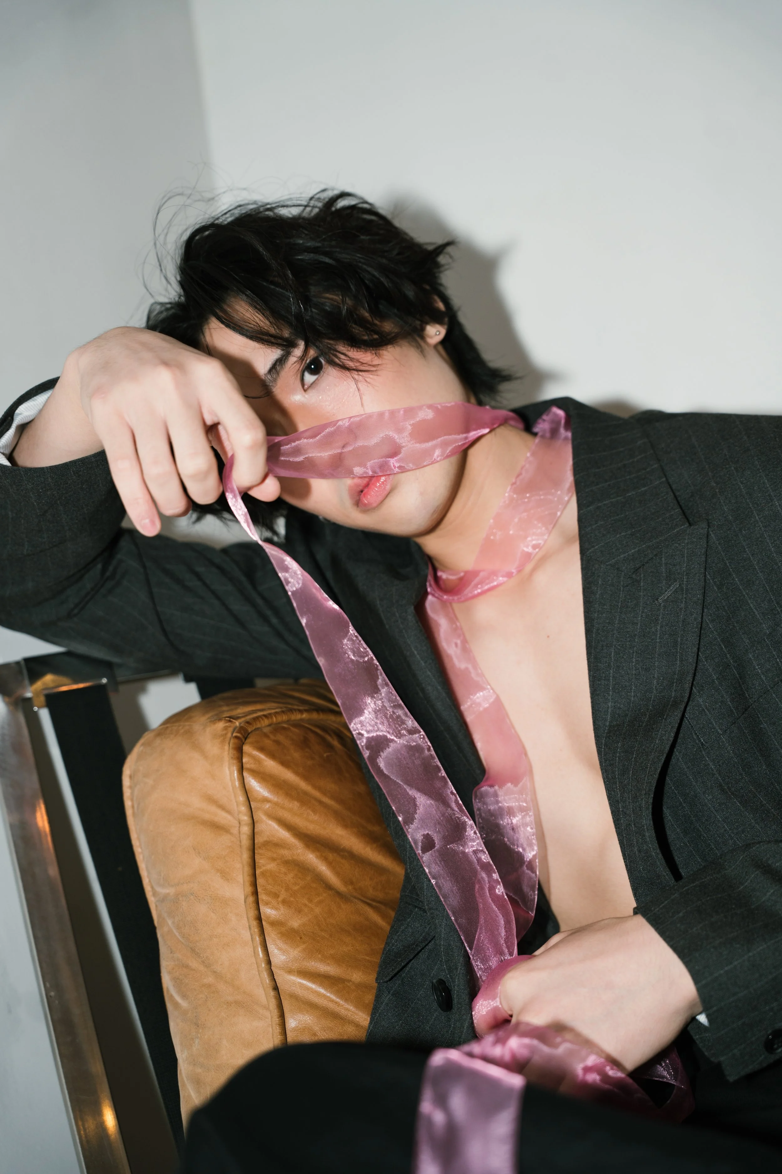 A young man with black hair and earrings wearing a dark blazer, partially covering his face with pink satin ribbons, sitting on a brown leather chair.