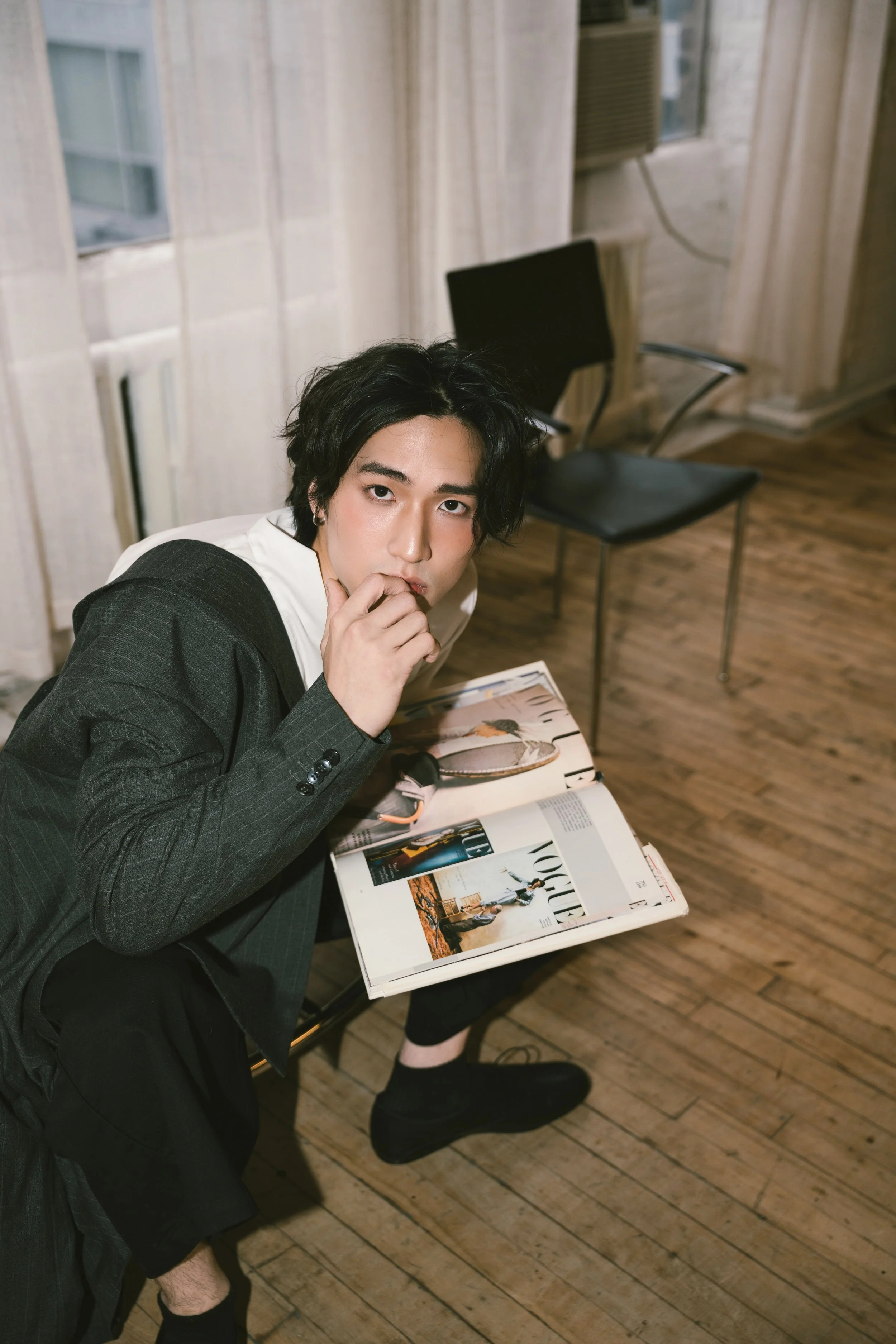 A young man with dark, wavy hair and fair skin, seated at a table with a magazine, in a room with wooden flooring, light-colored curtains, and a black chair in the background.