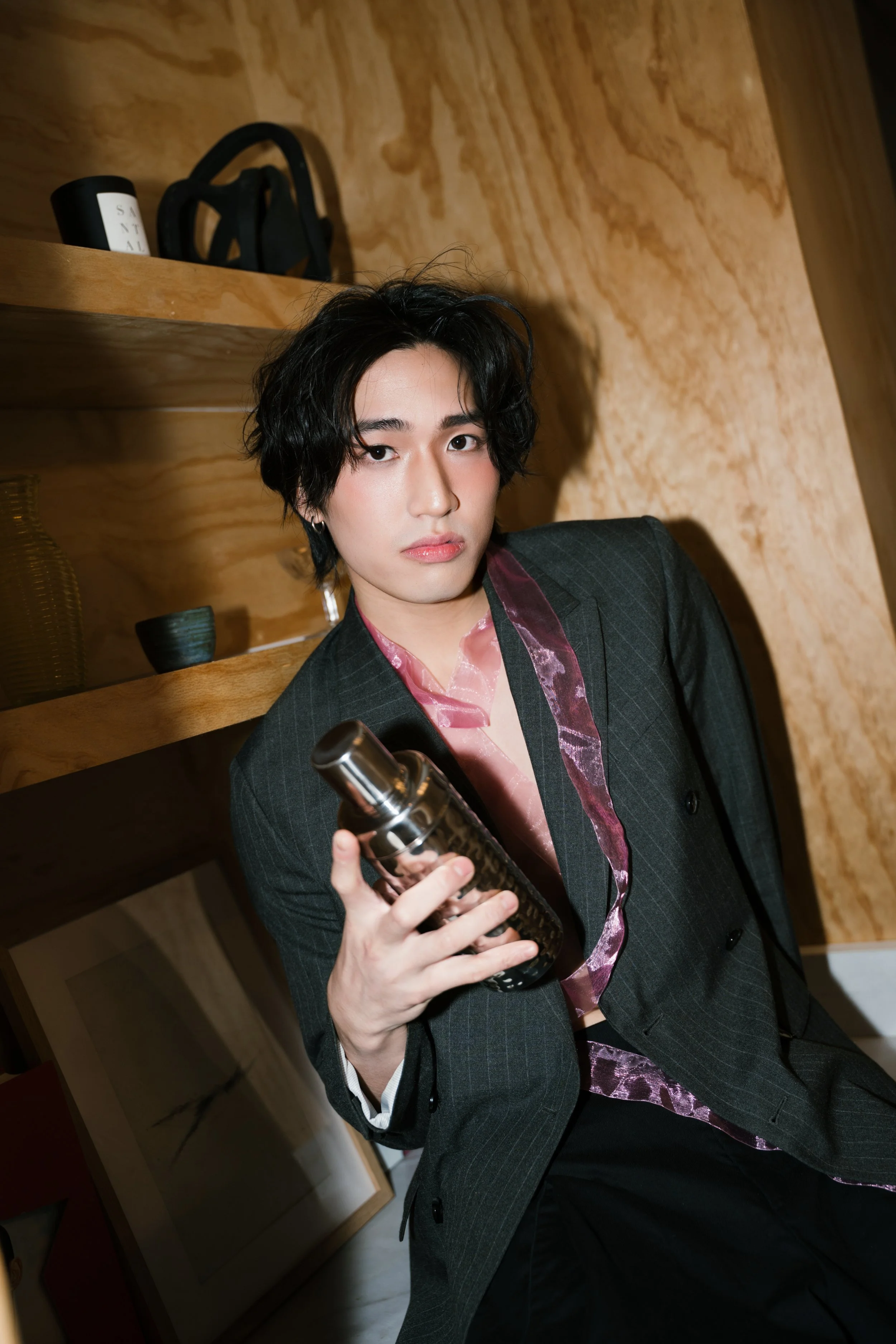 A young person with dark wavy hair, wearing a dark pinstripe suit and a pink satin shirt, holding a wine bottle, standing in a warmly lit room with wooden shelves and wall.
