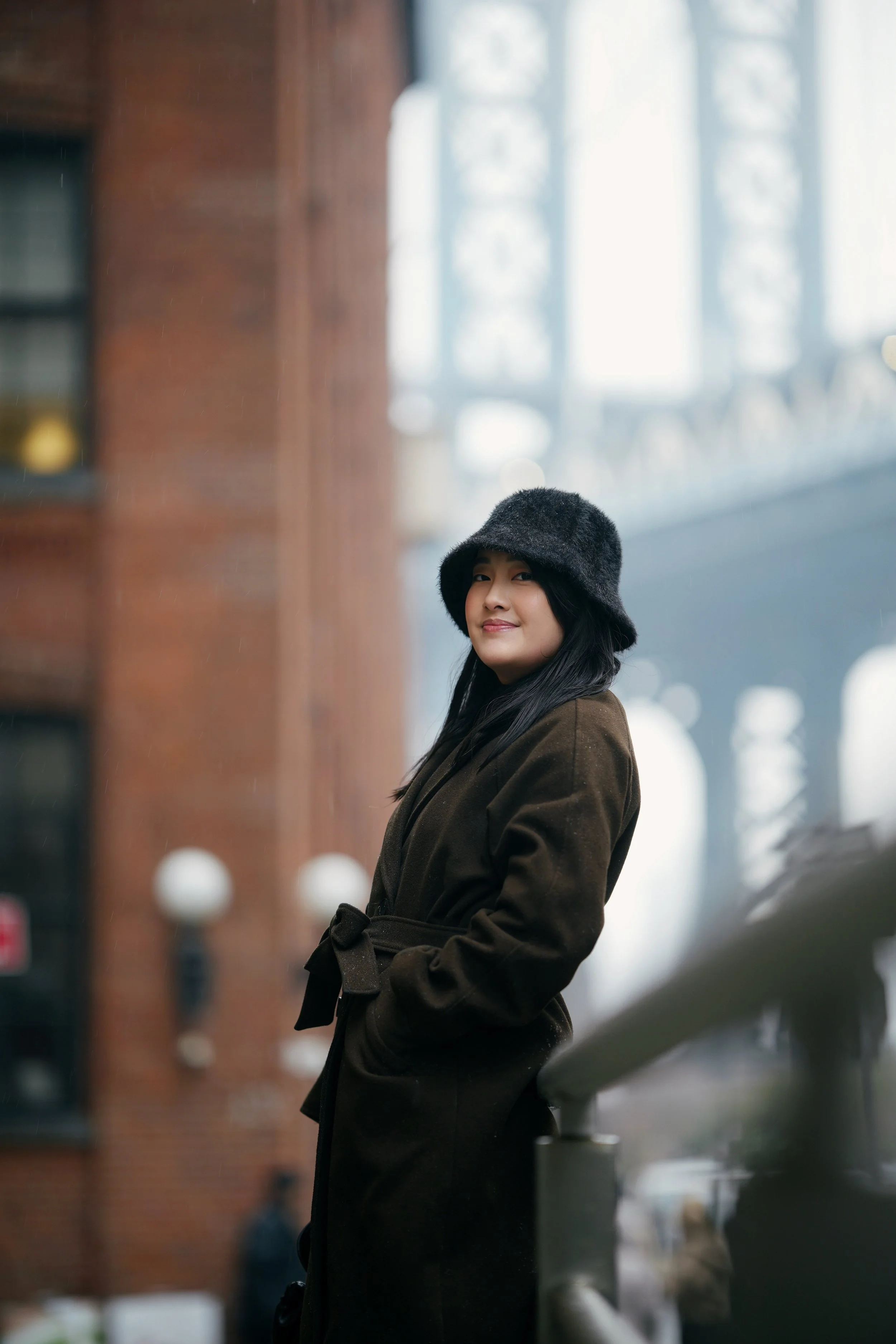 A woman with long black hair wearing a dark brown coat and a black fuzzy hat, smiling slightly, stands outdoors near a brick building with large windows in the background. She is partially turned to the side, with her hands in her coat pockets.
