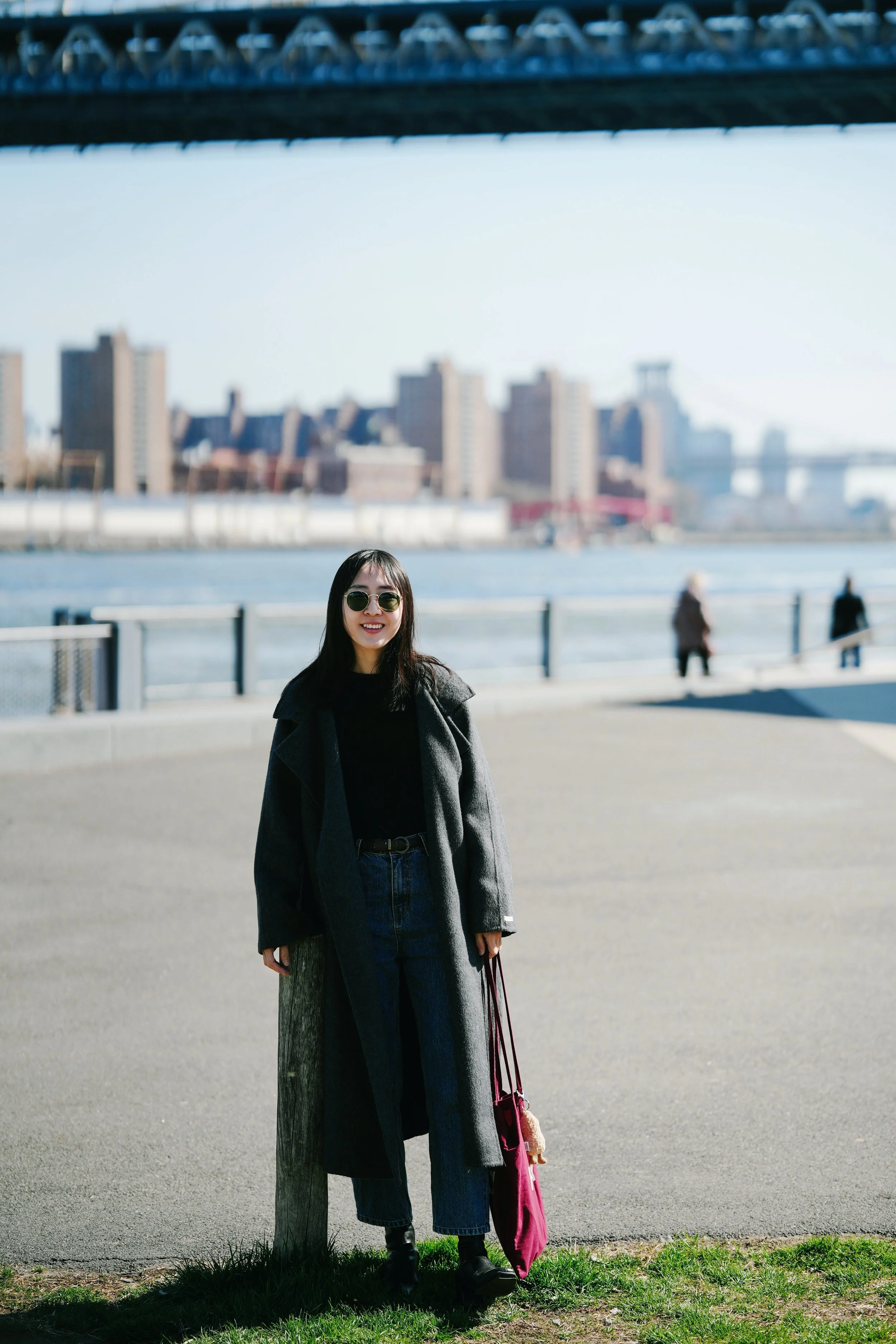 A woman with long dark hair and sunglasses smiling outdoors by a waterway with city buildings and a bridge in the background.