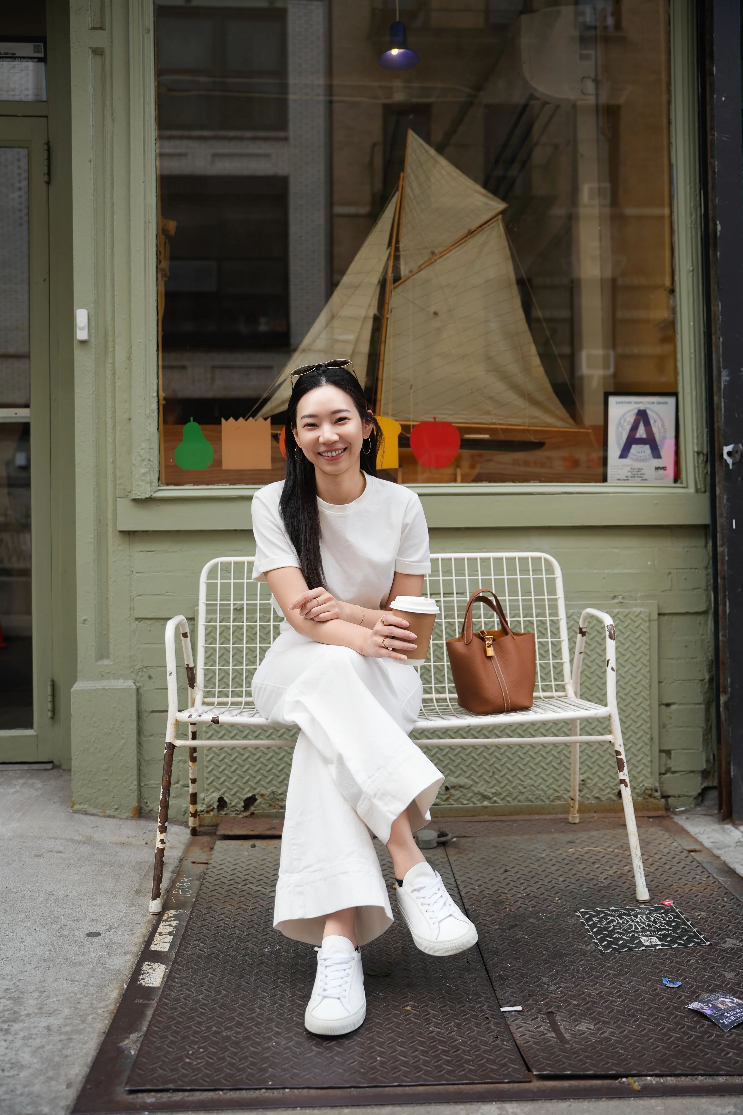 A young woman with long dark hair, wearing a white outfit and white sneakers, sitting on a white metal bench outside a storefront with a large glass window and green trim. She is smiling, holding a coffee cup, and has a brown handbag beside her. The 