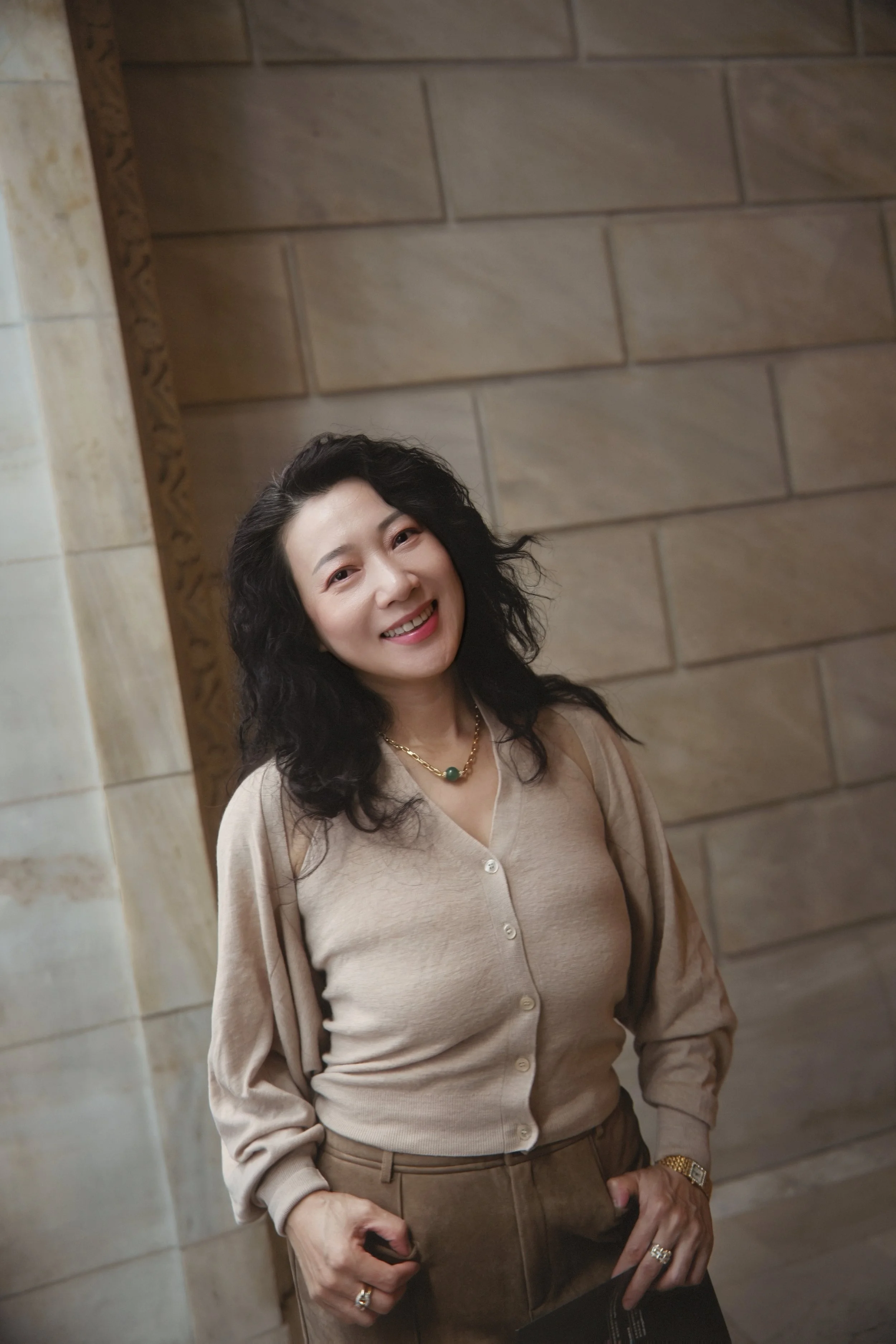 A smiling woman with black curly hair wearing a beige cardigan, a gold necklace with a green pendant, gold watch, and rings, standing against a stone wall.