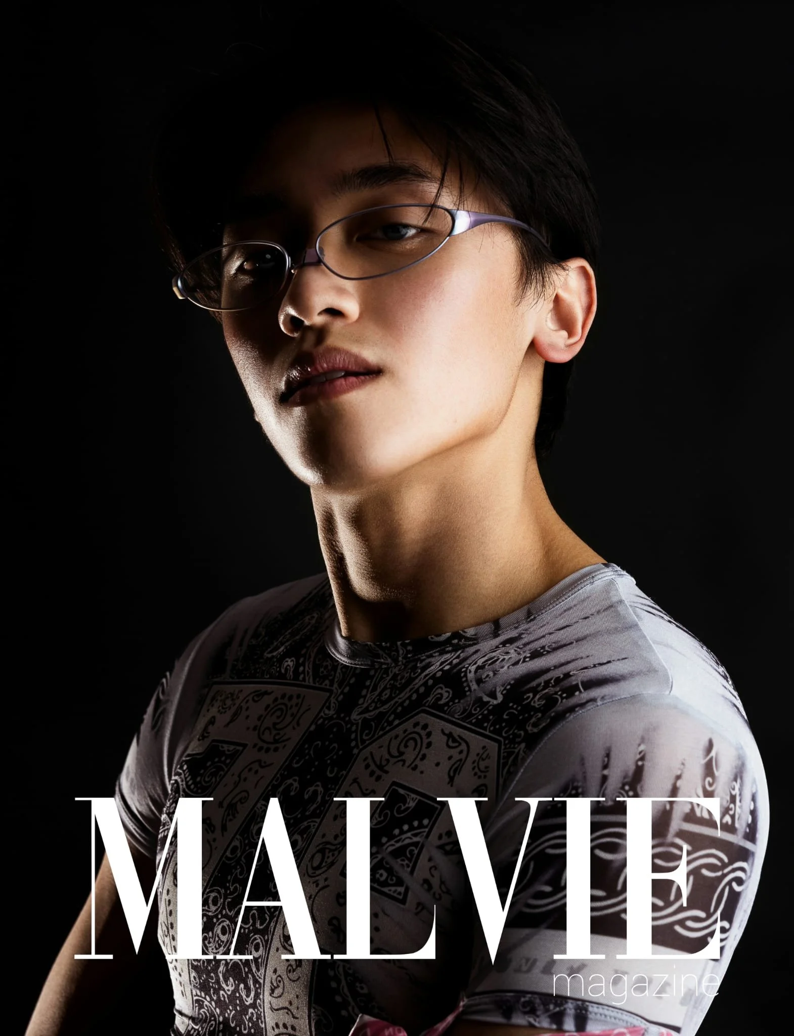 A young person with short dark hair, glasses, and a confident expression, wearing a patterned shirt, poses against a black background with the words "MAY WELL" and "magazine" overlaid.