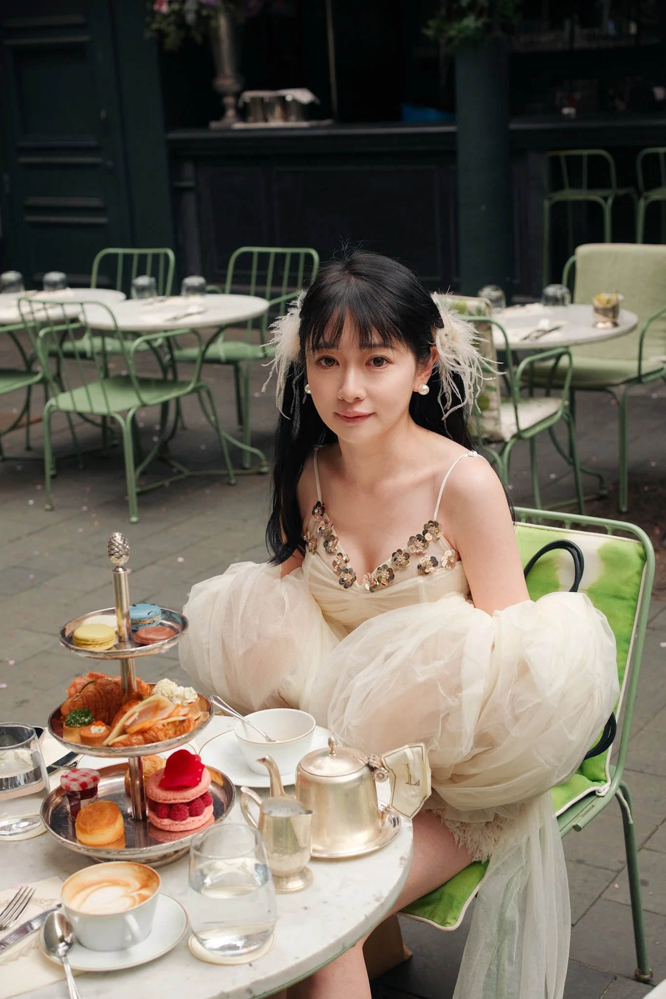 A woman with dark hair, wearing pearl earrings and a fancy cream-colored dress with sequined details, sitting at a table with a three-tiered tray of assorted sweets, tea, and coffee, in an outdoor café setting with green chairs and tables.