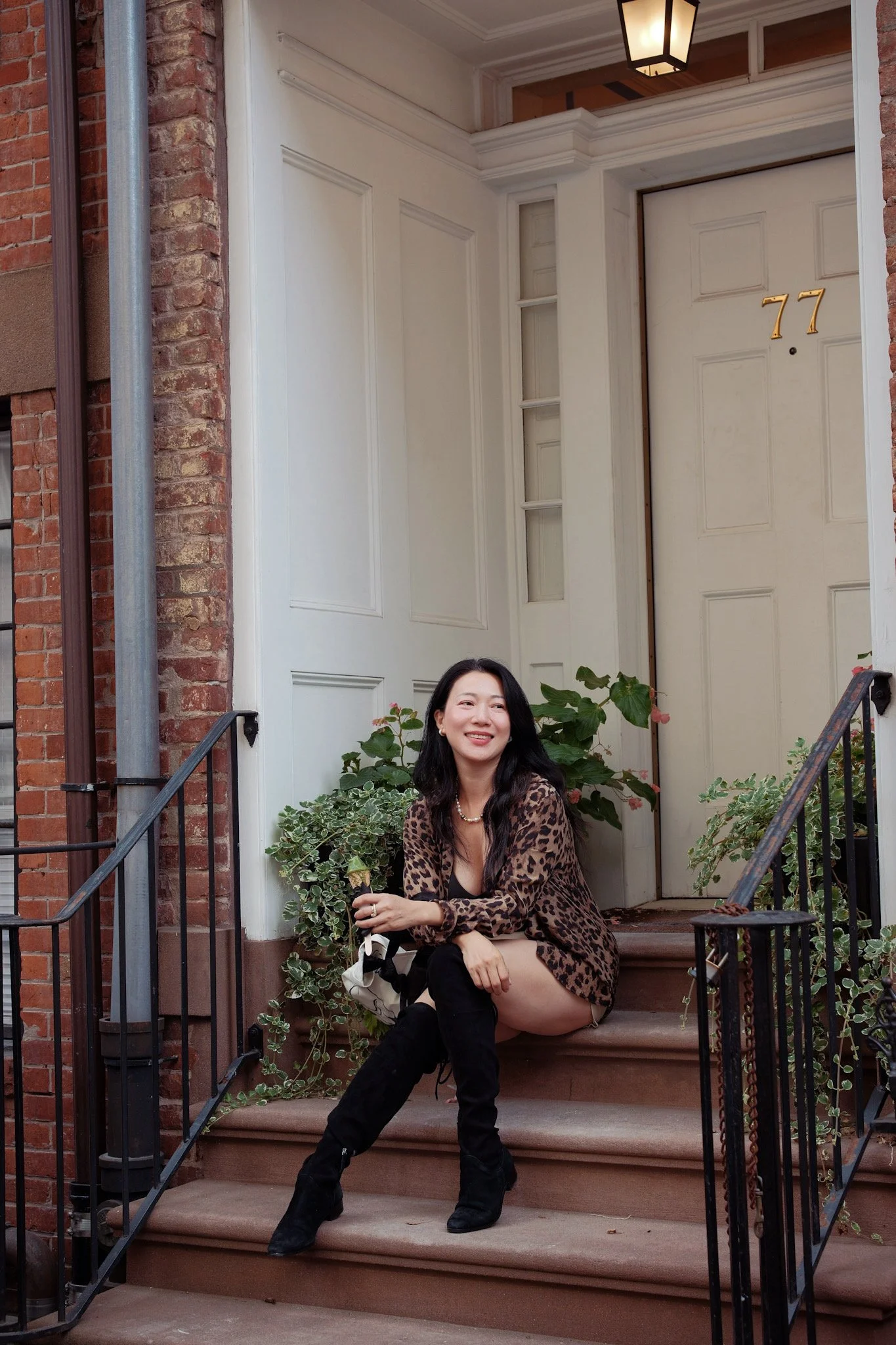 Young woman sitting on front steps of a brick house, smiling, wearing a leopard print dress and black boots, surrounded by plants and greenery.
