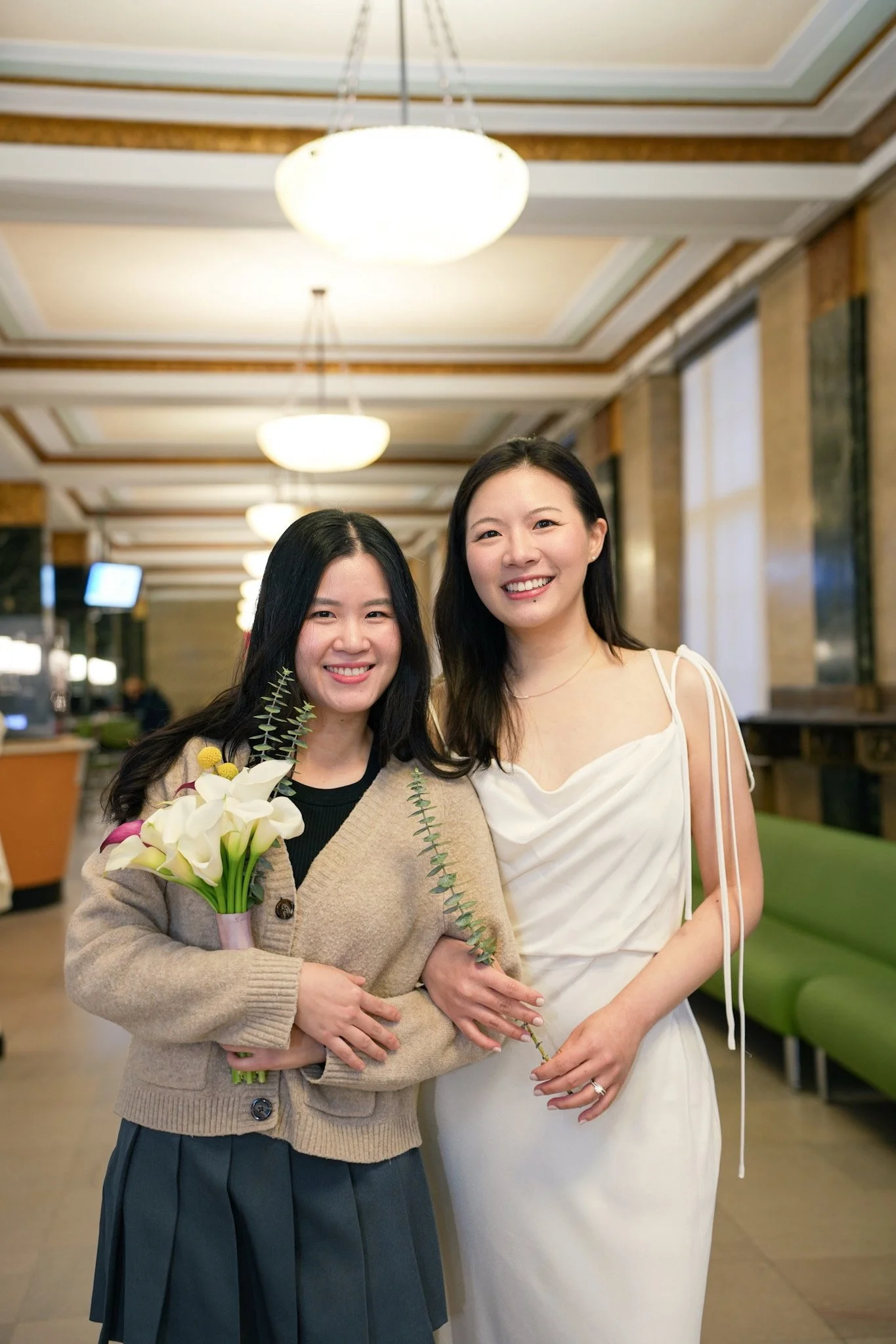 Two smiling women standing in a brightly lit indoor space, one holding a bouquet of flowers and both women with dark hair and light skin.