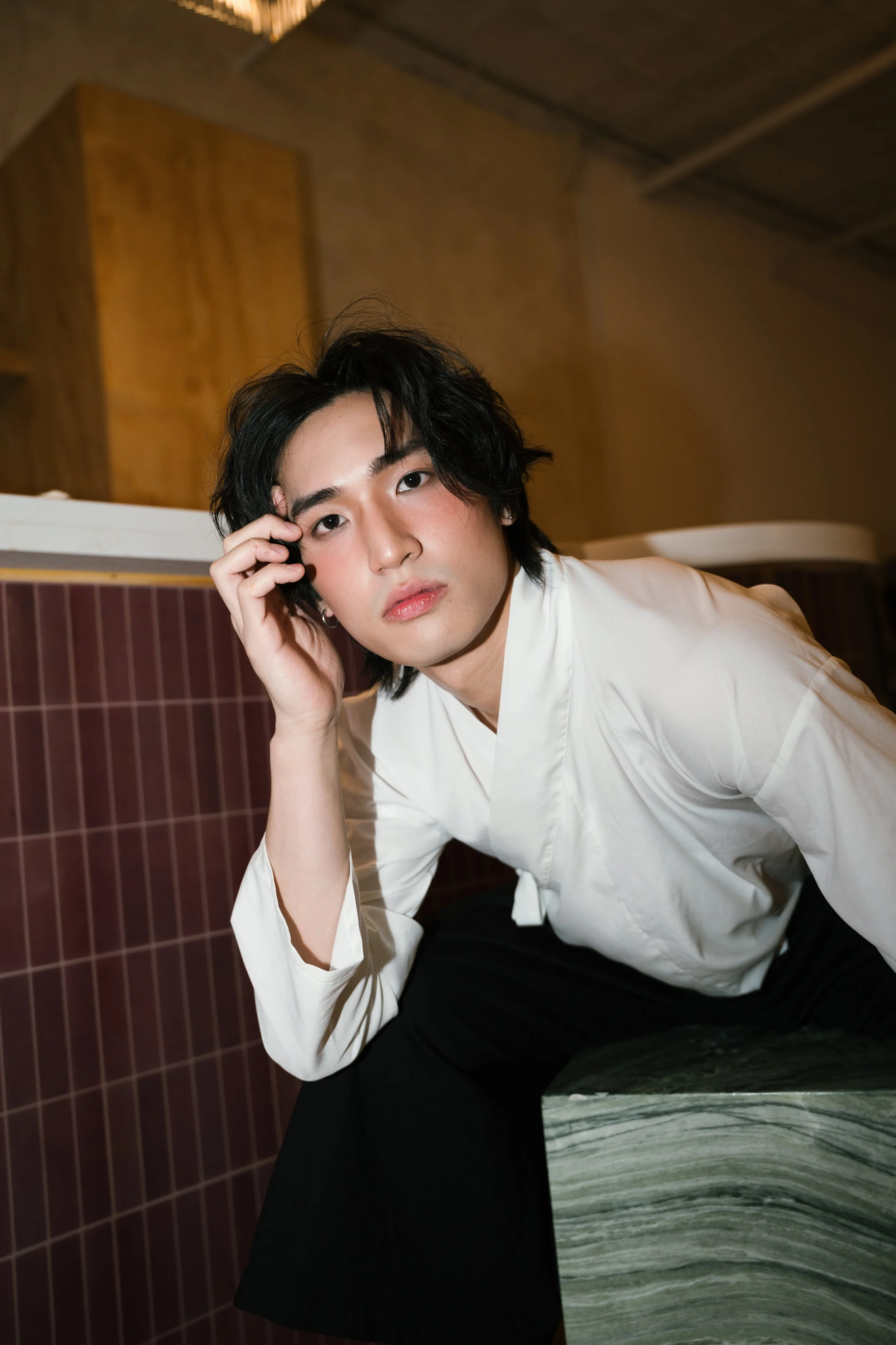 A young man with black wavy hair and fair skin, wearing a white shirt, is sitting on a black surface in a room with brown tile walls. He has a relaxed expression and is touching his face with his left hand, looking directly at the camera.
