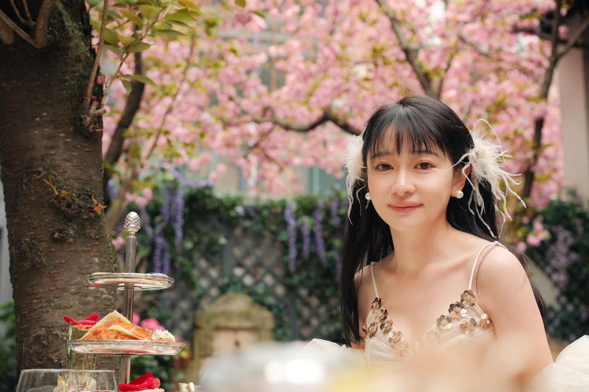 Young woman with black hair and pearl earrings standing outdoors under pink cherry blossom trees, wearing a light-colored dress with floral sequins, with a tiered stand of assorted small desserts in front of her.