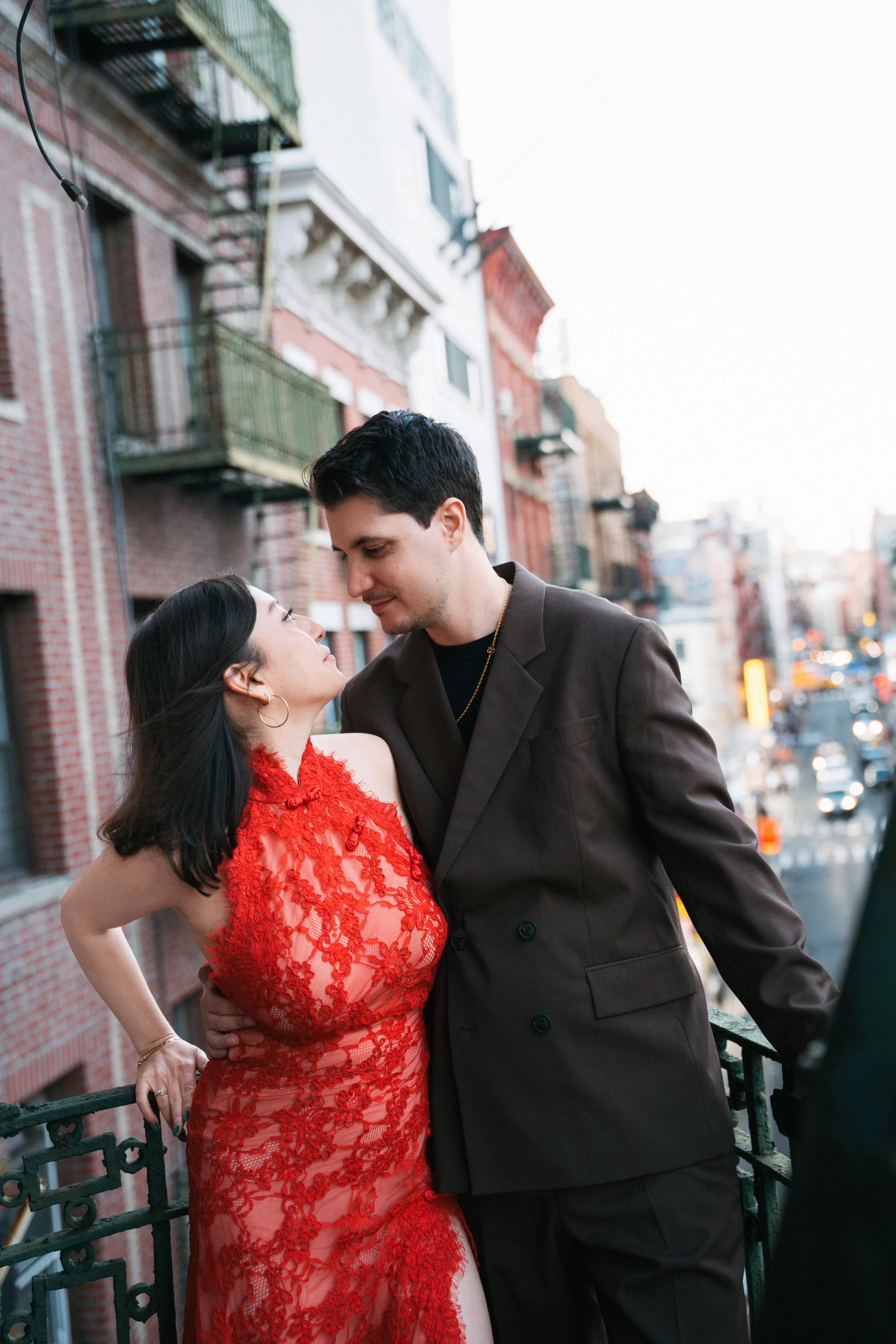 A man and a woman are close together on a city balcony, looking into each other's eyes. The woman wears a red lace dress, and the man is in a dark blazer. The background shows a city street with buildings, cars, and a bright sky.