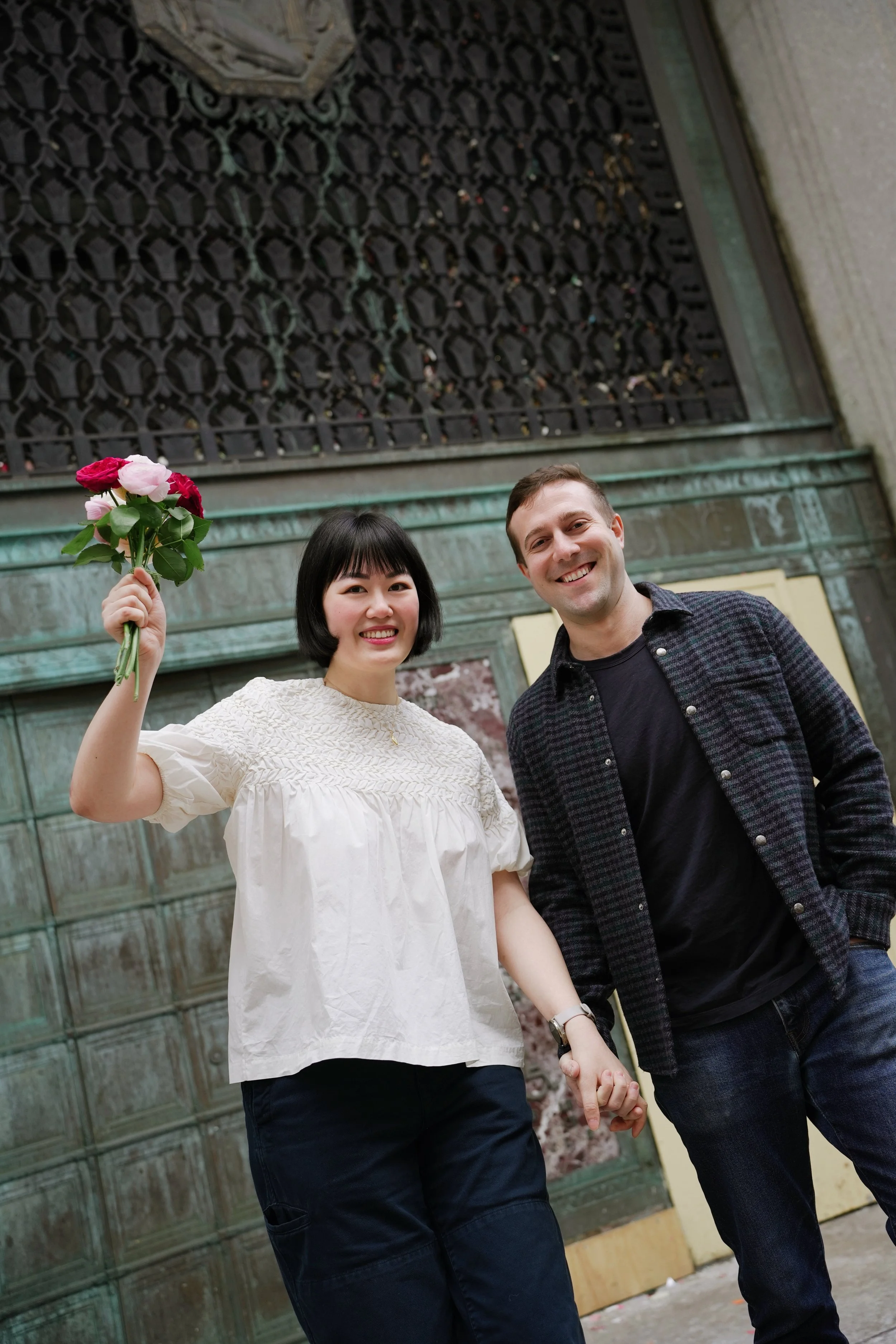 A woman holding a bouquet of pink and red roses and a man smiling, holding hands, standing outside in front of a decorative wall.
