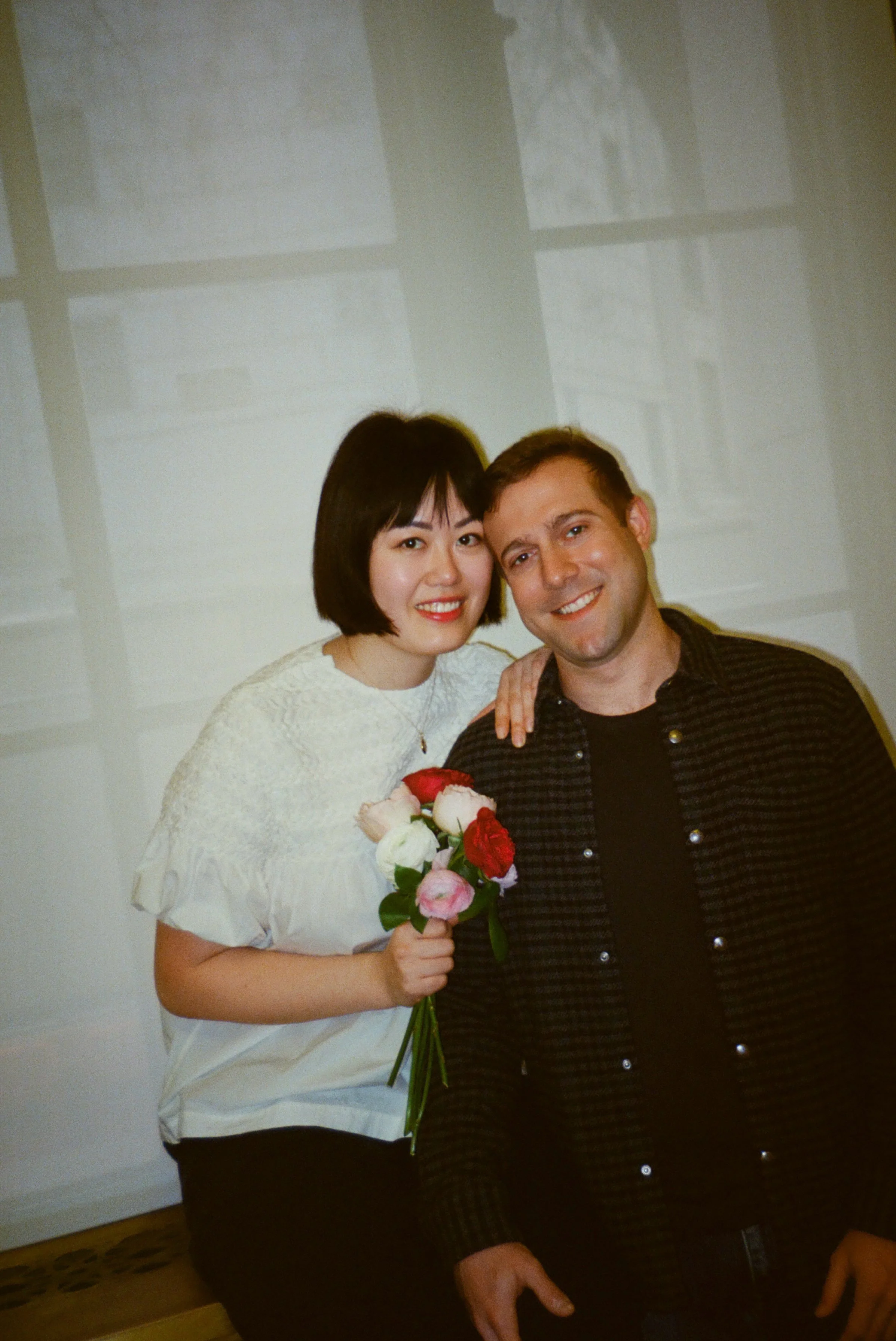 A woman with short dark hair holding a bouquet of pink and white roses, standing next to a man with short brown hair, both smiling and close together indoors.