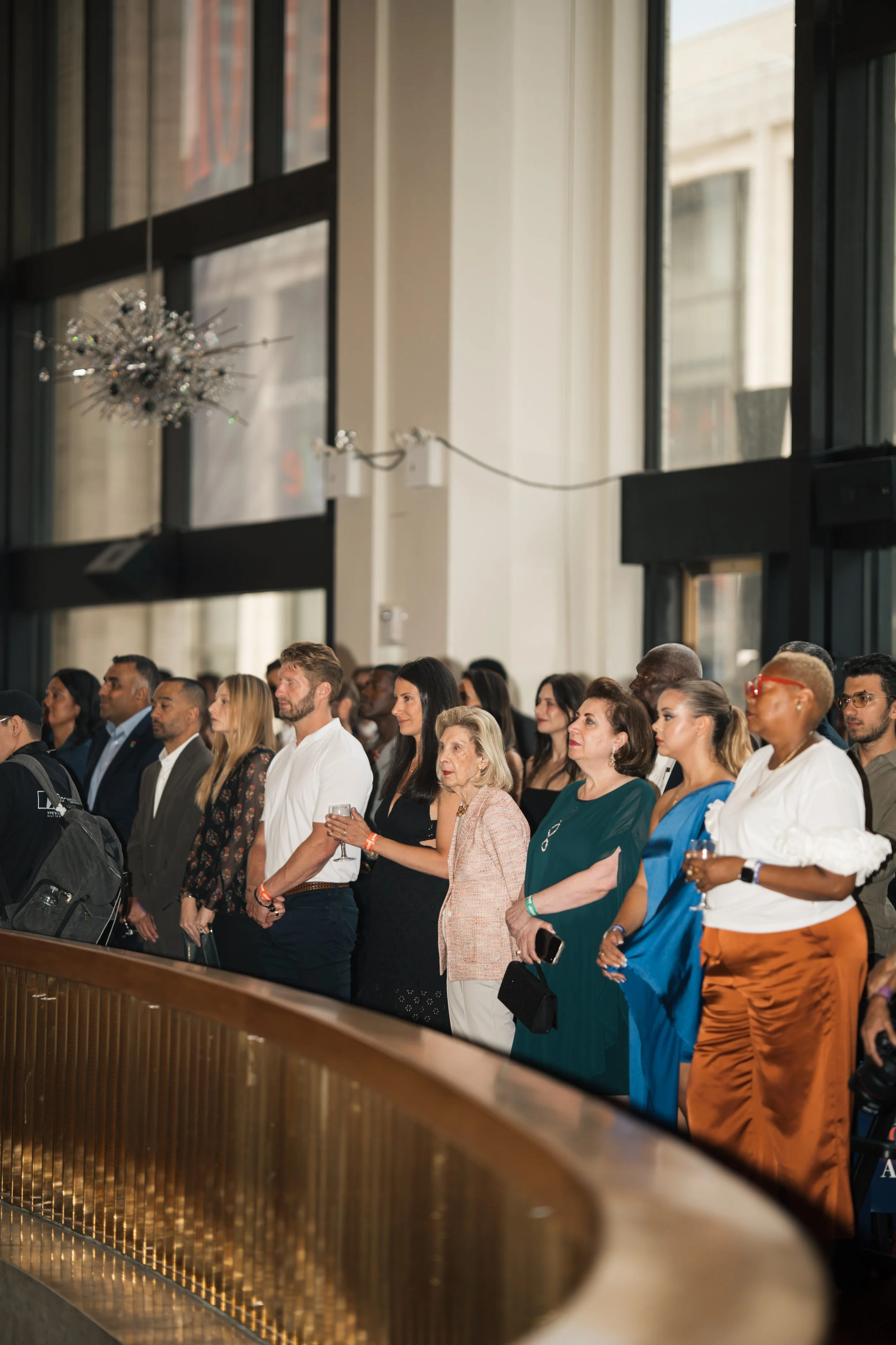 A diverse group of people attending an event, standing near a wooden railing inside a building with large windows and a decorative chandelier.