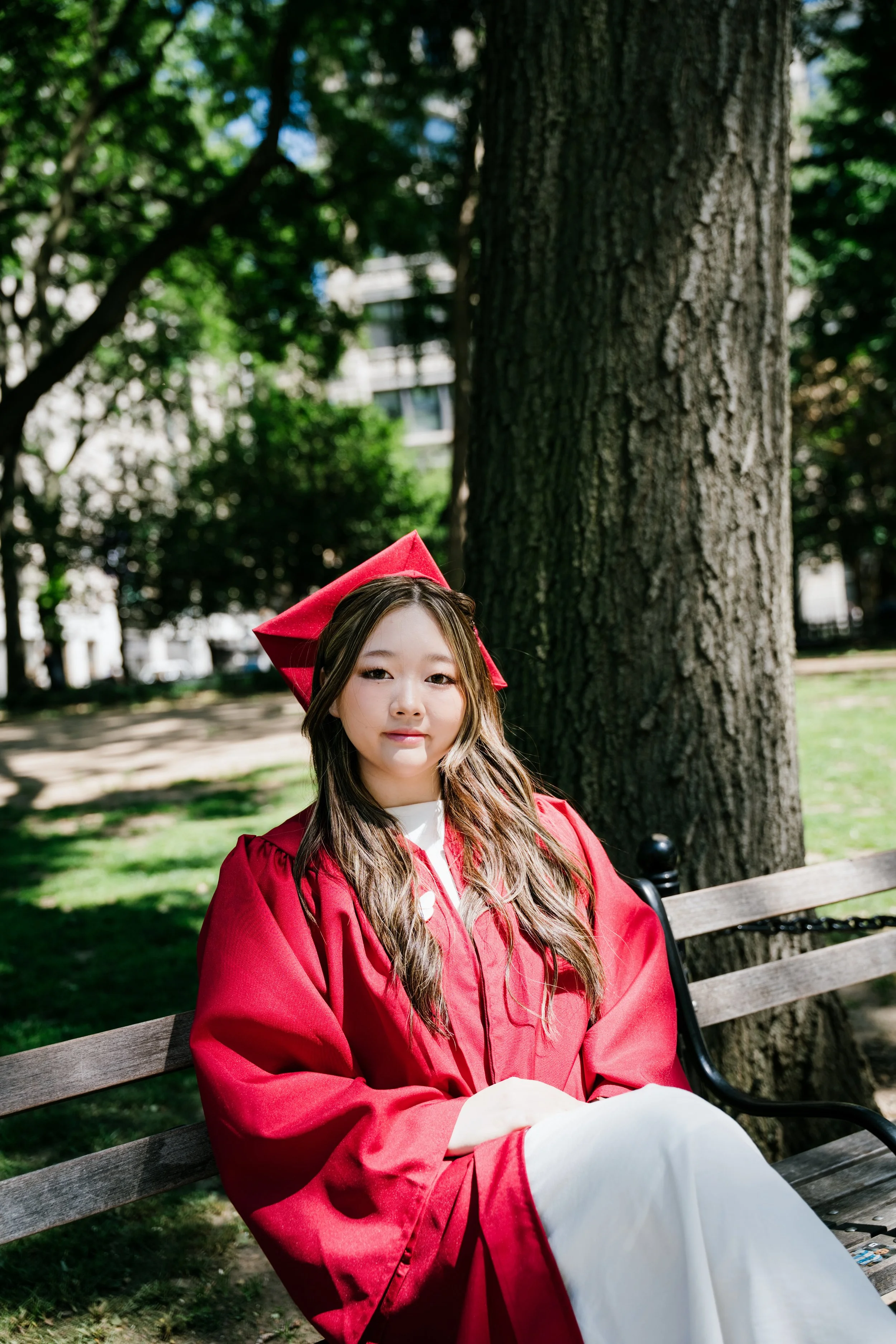Young woman in a red graduation gown and cap sitting on a park bench under a large tree, with greenery and a building in the background.