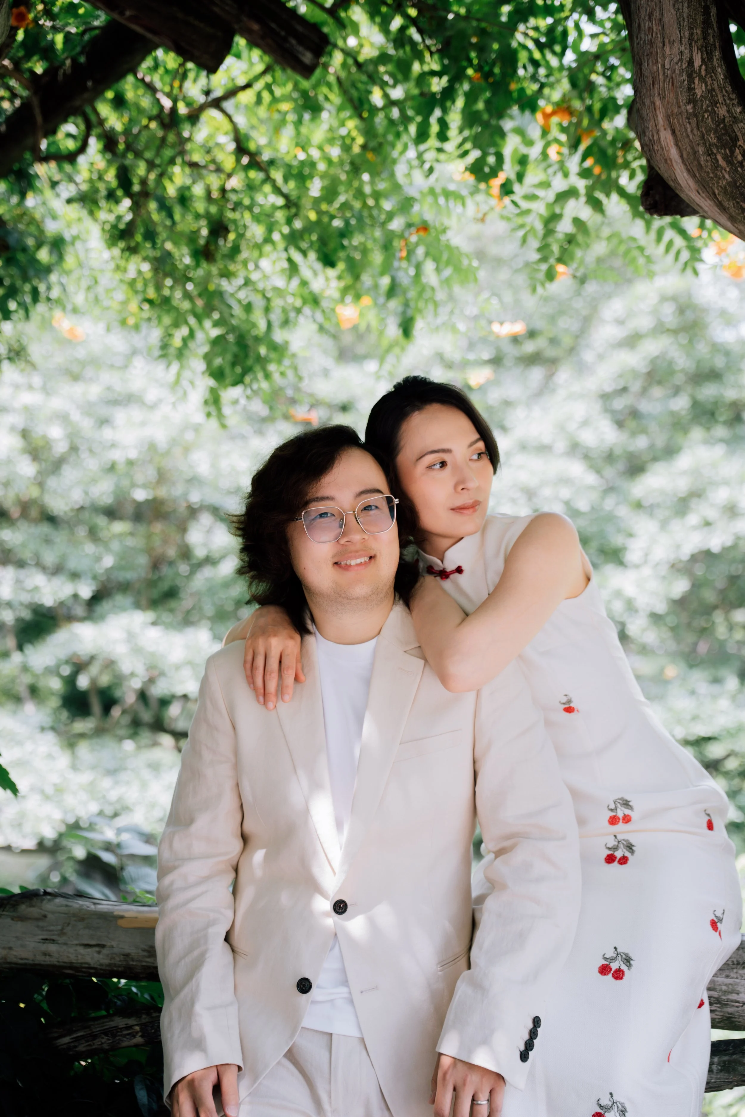 A man and a woman are outside under a tree. The man is wearing a cream-colored suit and glasses, and the woman is hugging him from behind, wearing a white dress with embroidered cherries. They are smiling and looking into the distance.
